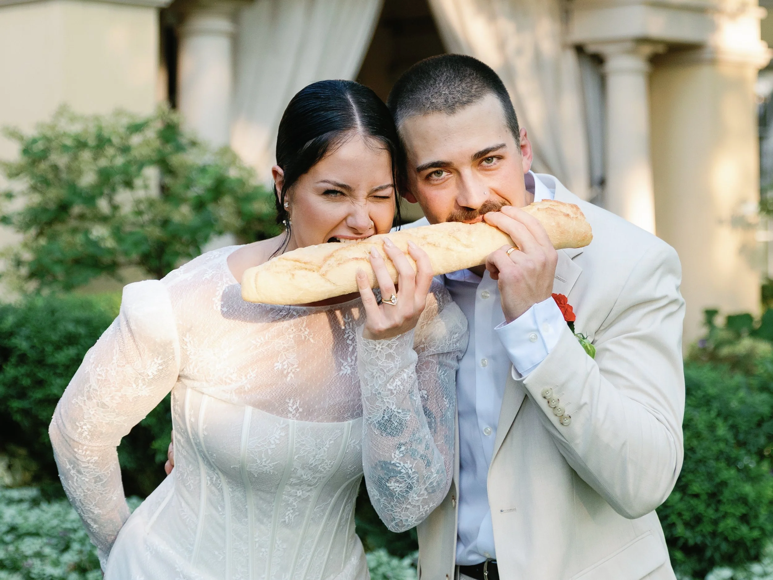 Couple in wedding attire sharing a giant baguette outdoors with greenery and columns in the background.
