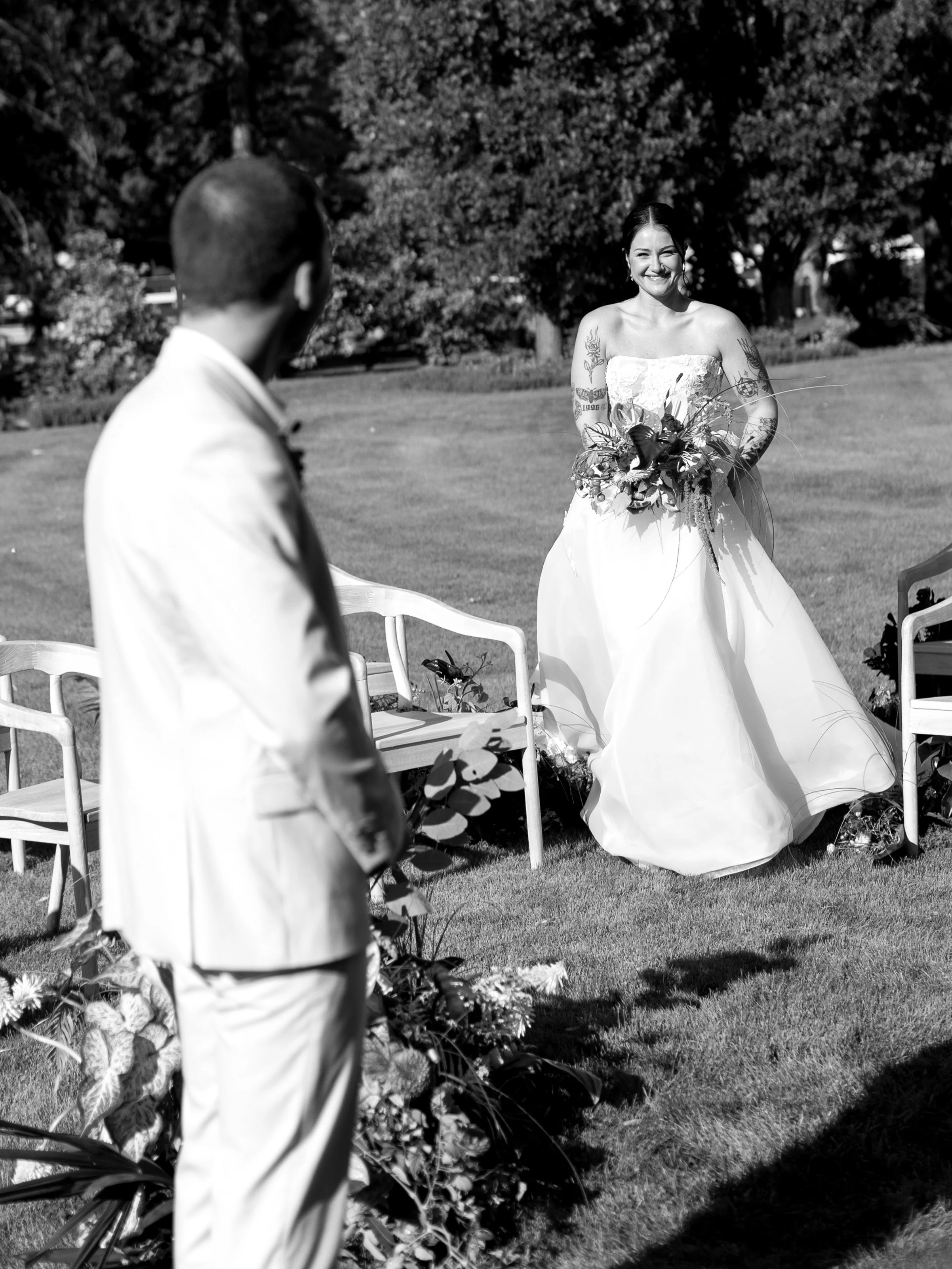 A bride in a strapless wedding dress holding a bouquet of flowers looks at a groom in a light-colored suit during an outdoor wedding ceremony on a grassy area with trees in the background.