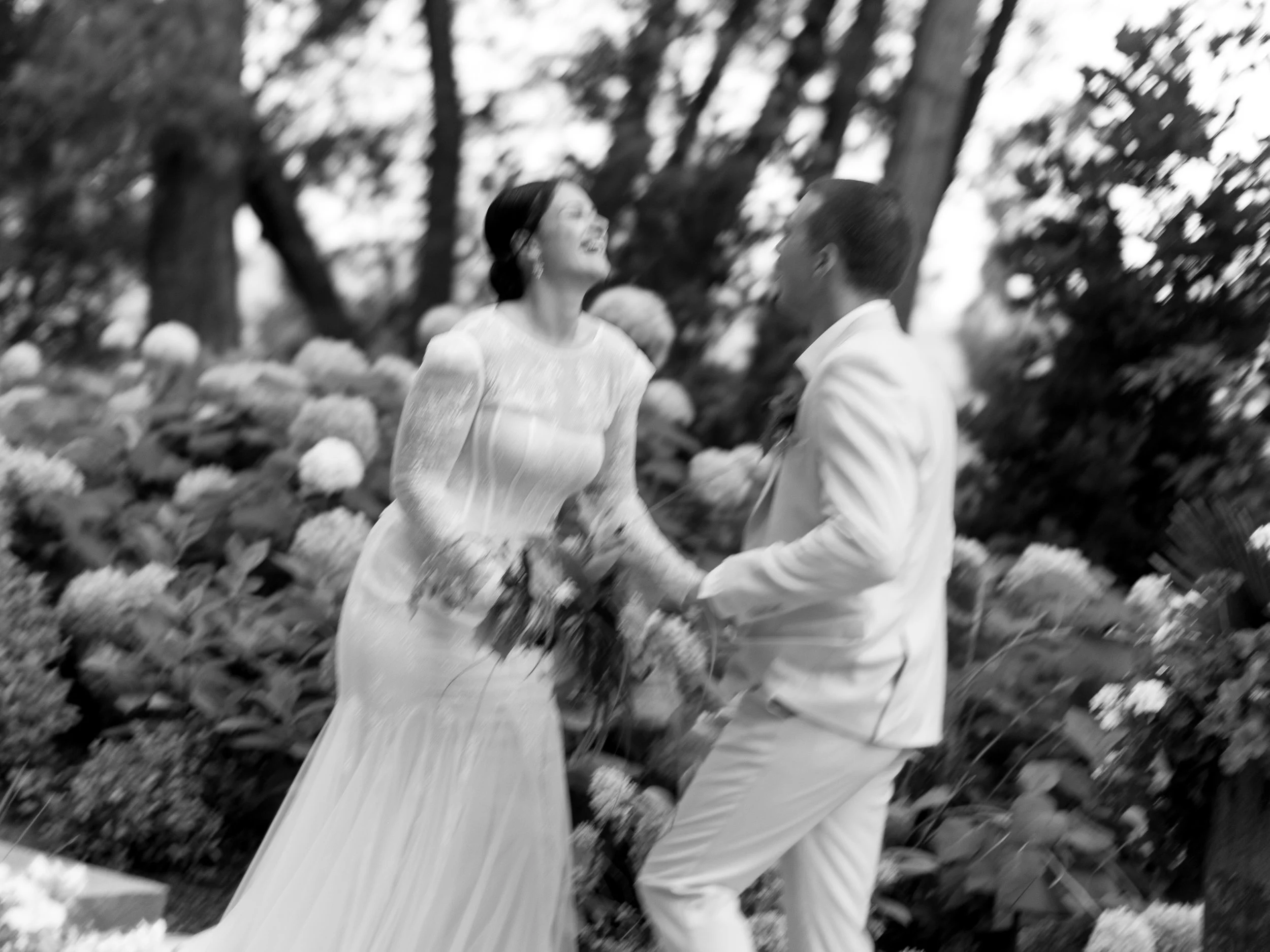 A bride and groom holding hands and smiling at each other outdoors, surrounded by flowers and trees.