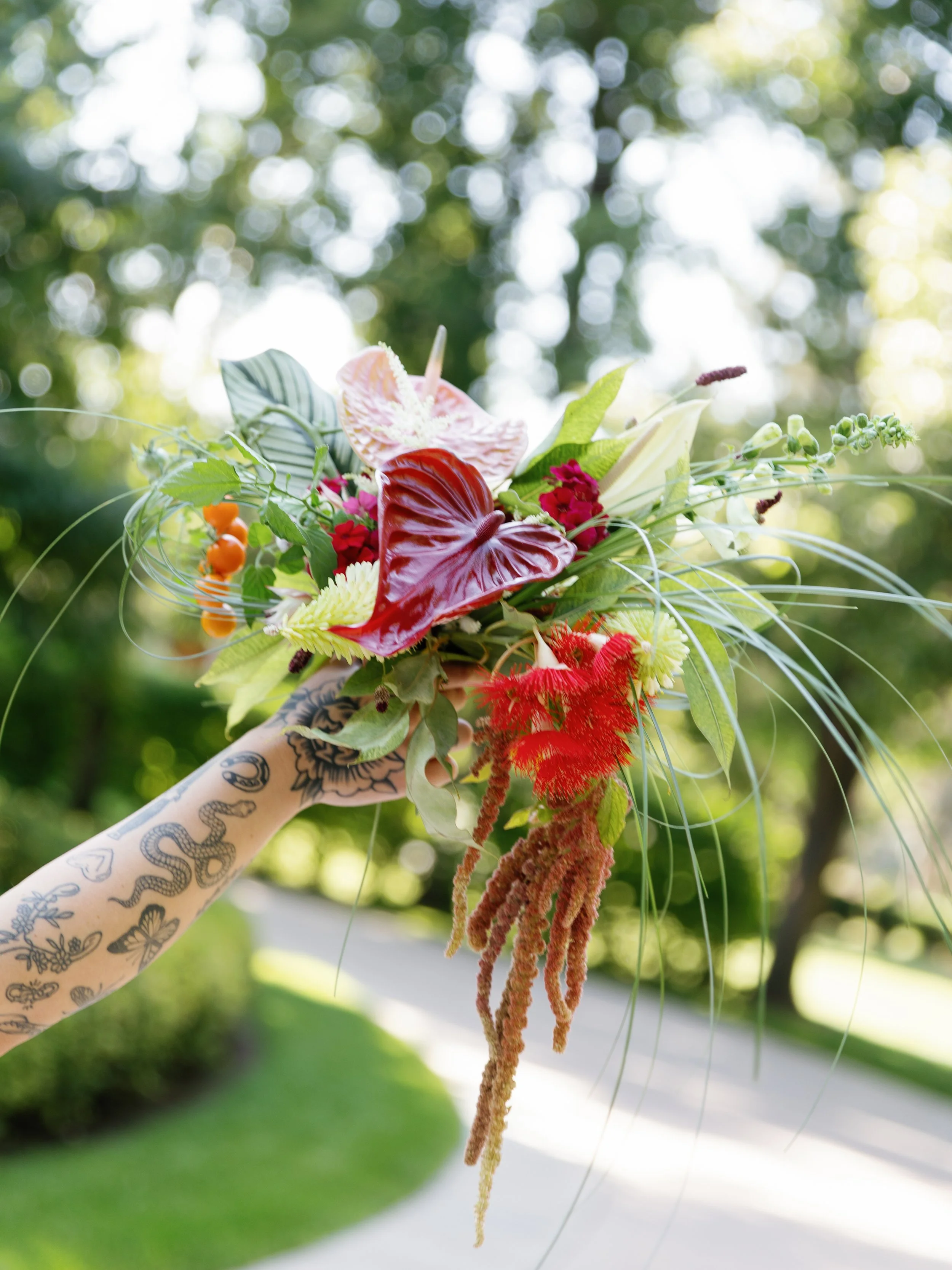 Person's arm with tattoos holding a colorful bouquet of flowers and greenery outdoors with trees and sunlight in the background.
