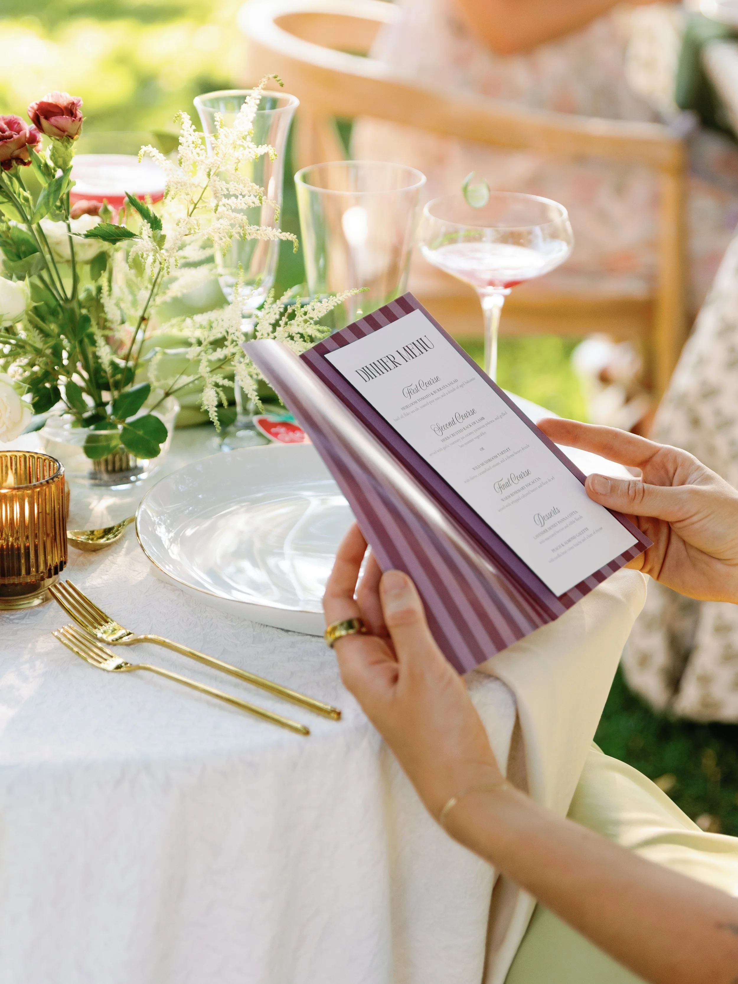Person holding a restaurant dinner menu at a table set with flowers, glasses, and gold utensils.