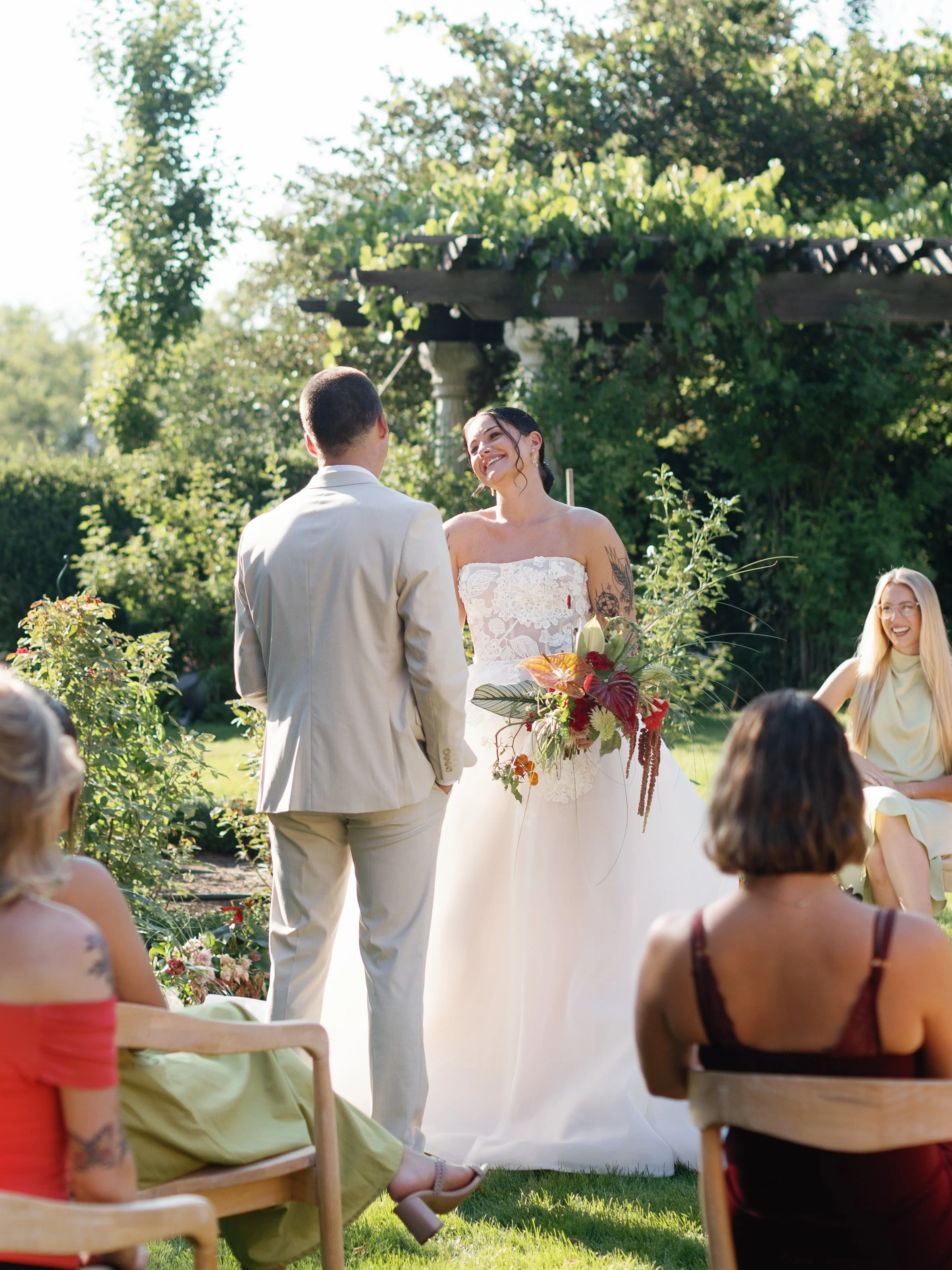 A outdoor wedding ceremony with a bride and groom facing each other, surrounded by seated guests. The bride is holding a bouquet and smiling, wearing a strapless white wedding dress. The groom is dressed in a light-colored suit. The scene is set in a