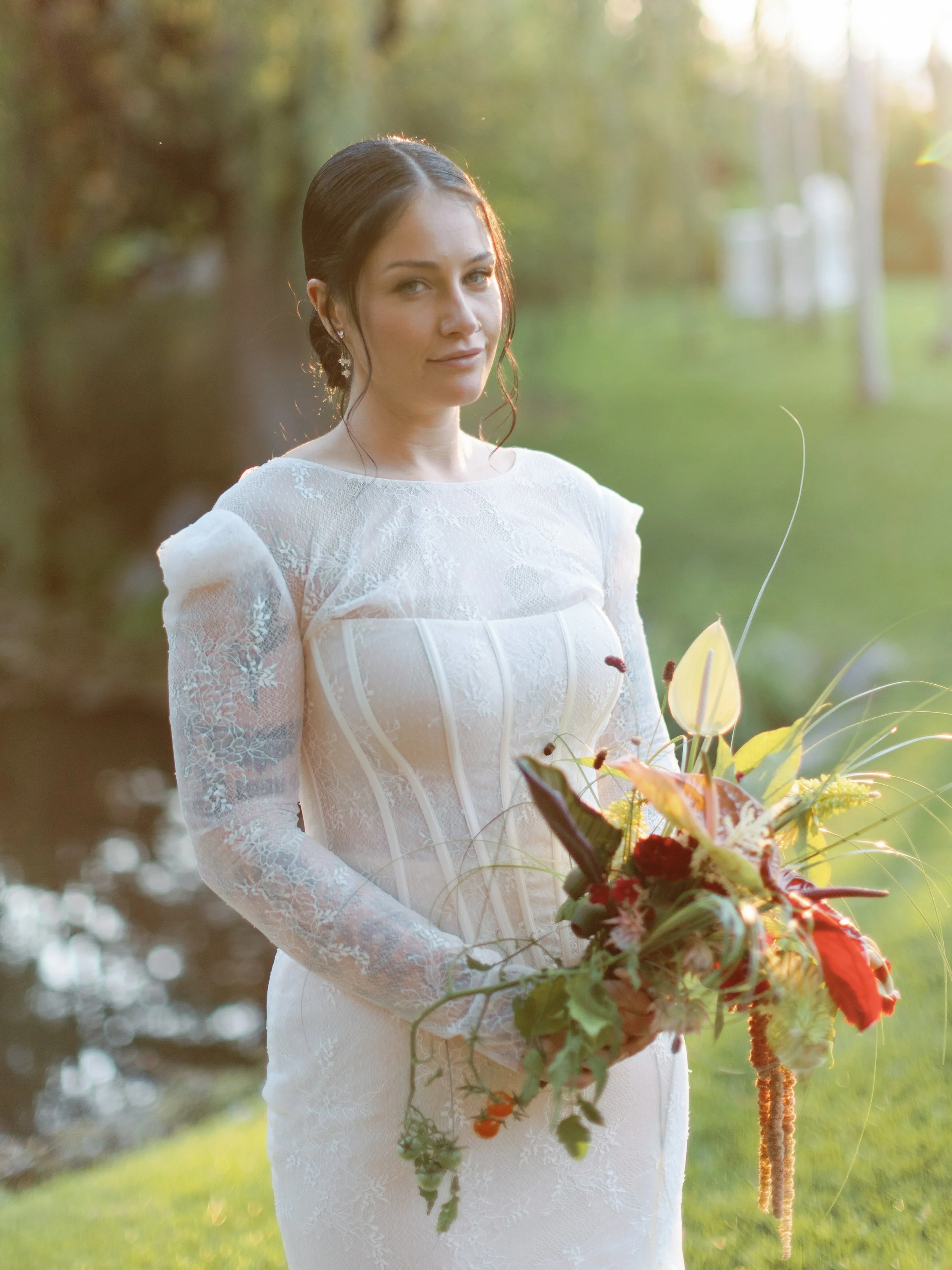 A woman in a white lace wedding dress standing outdoors near a pond, holding a bouquet of flowers, with trees and sunlight in the background.