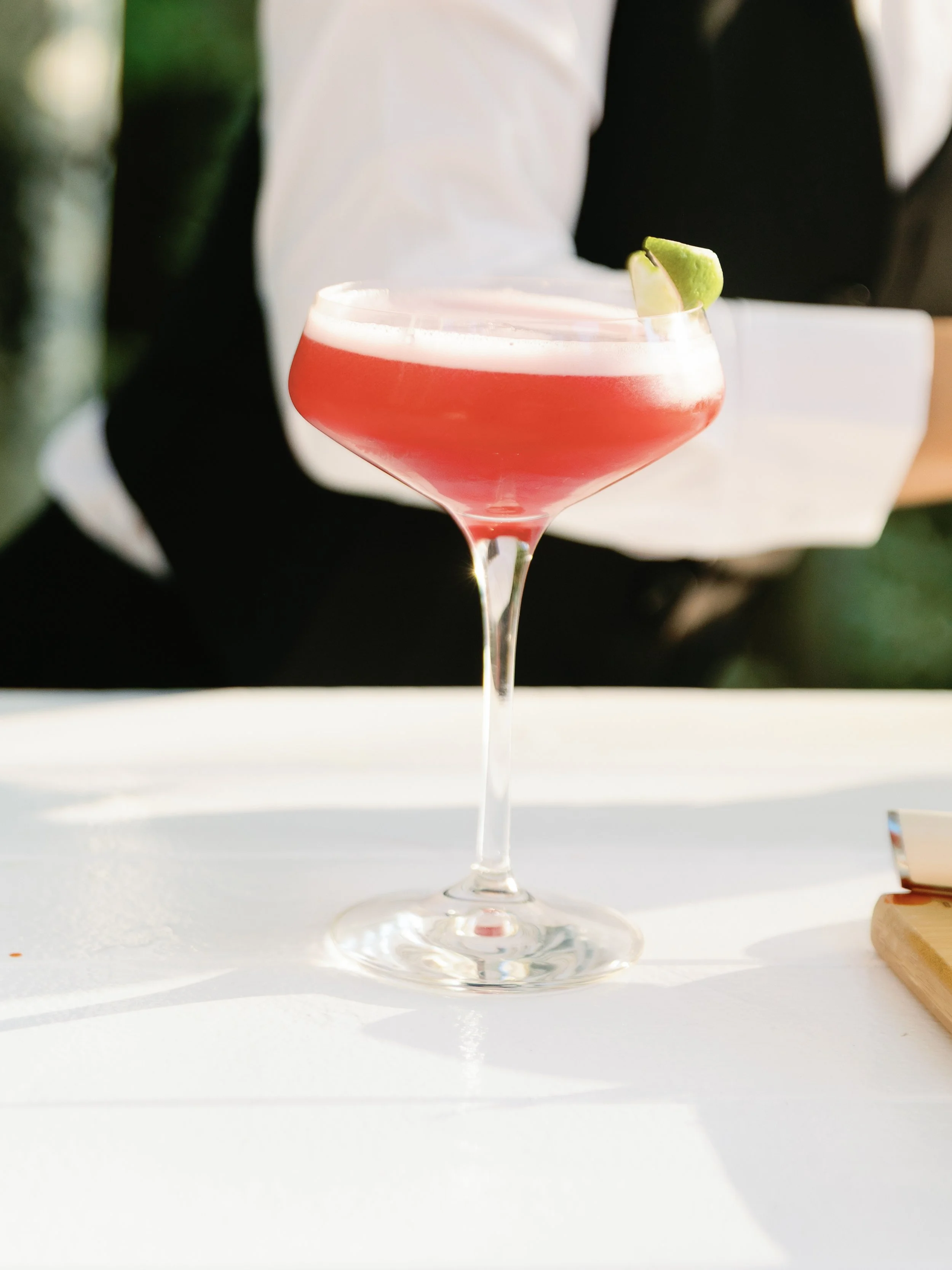 A pink cocktail with a lime wedge on the rim served in a coupe glass on a white table.