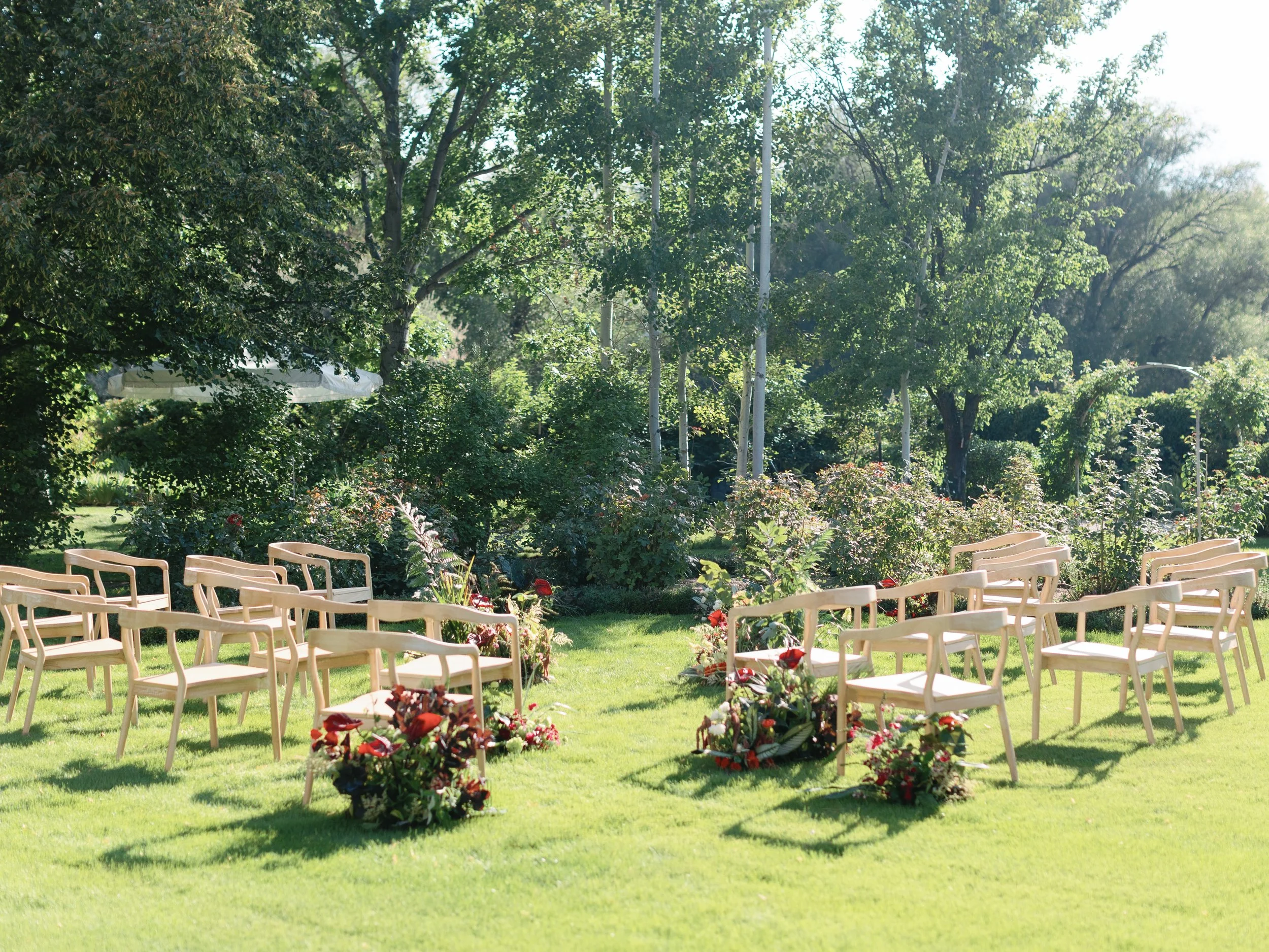 Outdoor wedding ceremony setup with wooden chairs arranged in two rows, decorated with floral arrangements, on a grassy lawn with trees in the background on a sunny day.