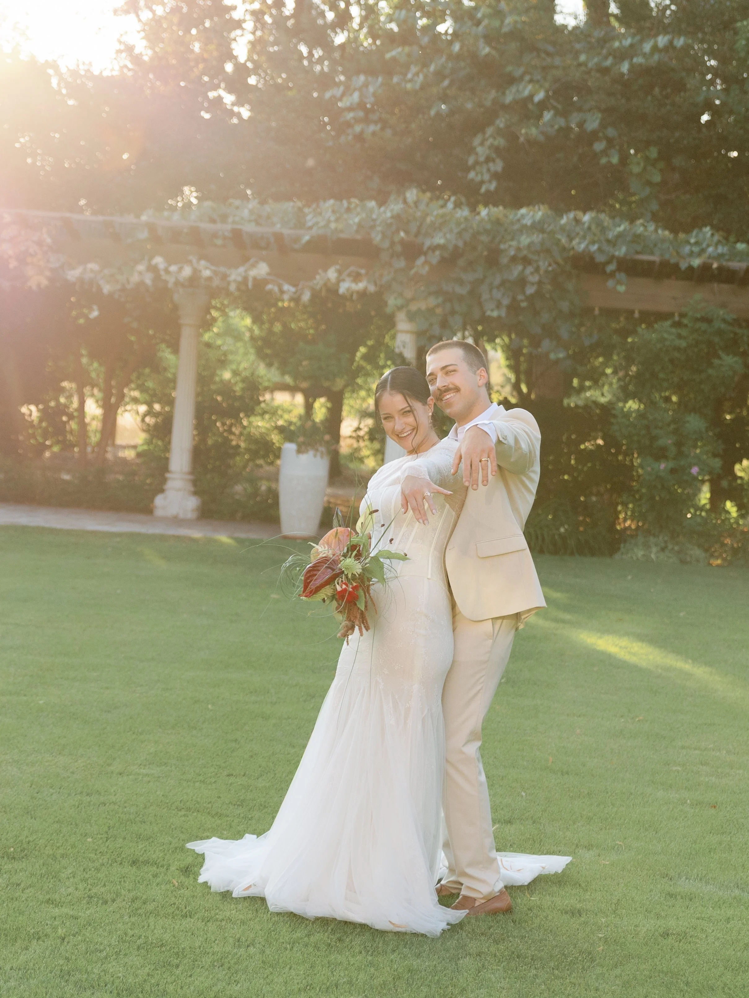 A newlywed couple poses outdoors on a sunny day, holding hands and smiling, with the bride wearing a white gown and the groom in a beige suit. The bride holds a bouquet of flowers, and they stand on a grassy lawn with trees and a pergola in the backg