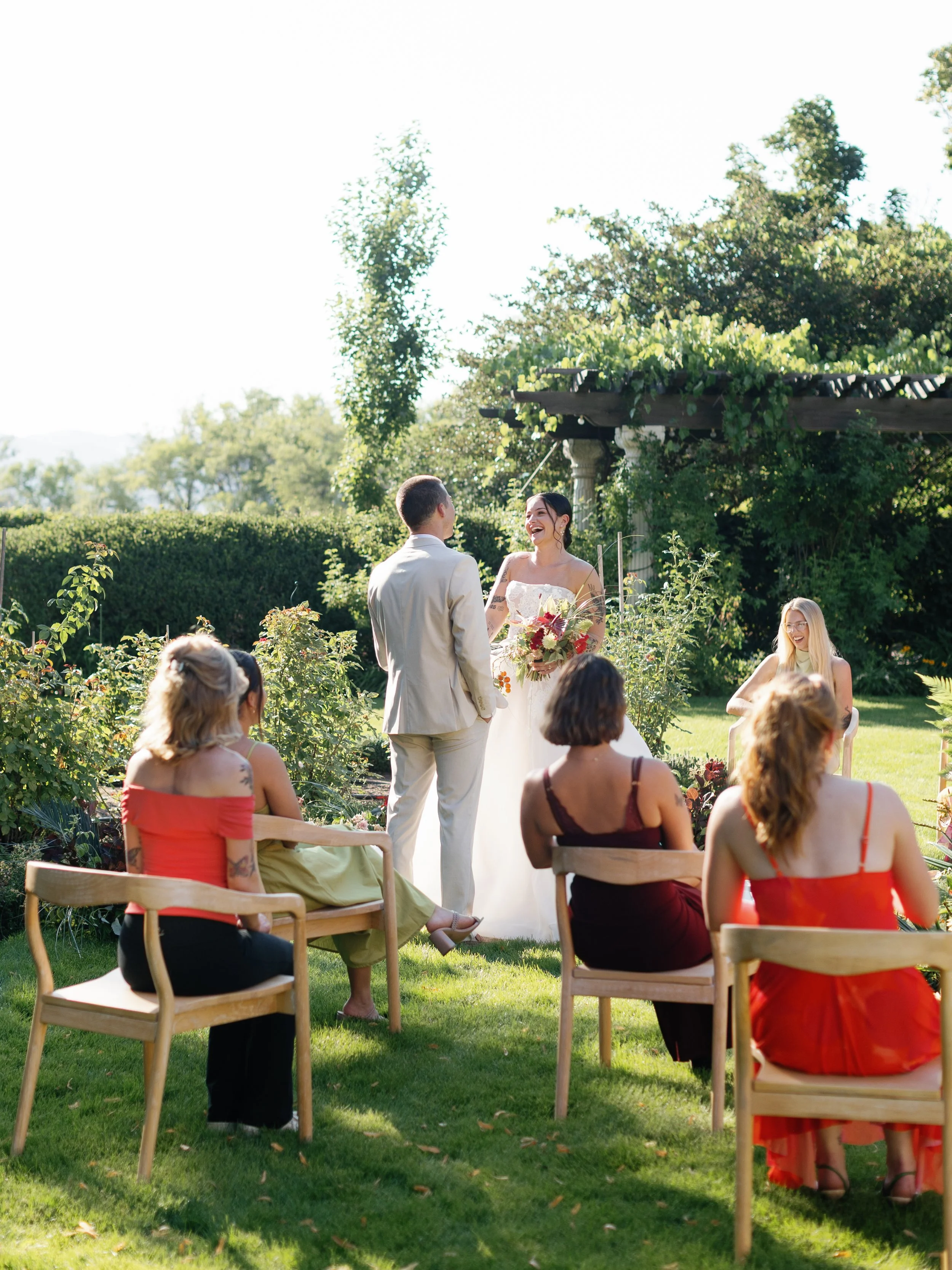 A couple getting married outdoors on a sunny day, standing in front of an officiant, with friends seated on wooden chairs watching the ceremony.