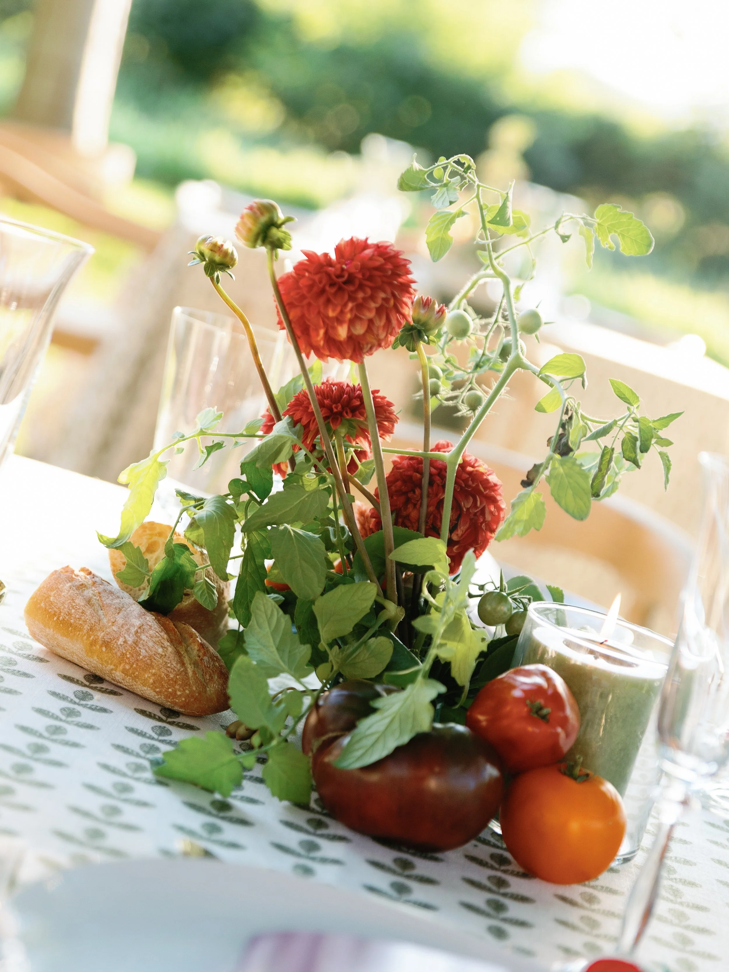 Table centerpiece with red dahlias, green leafy vine, tomatoes, a green candle, a loaf of bread, and glassware on a white tablecloth outdoors with sunlight.
