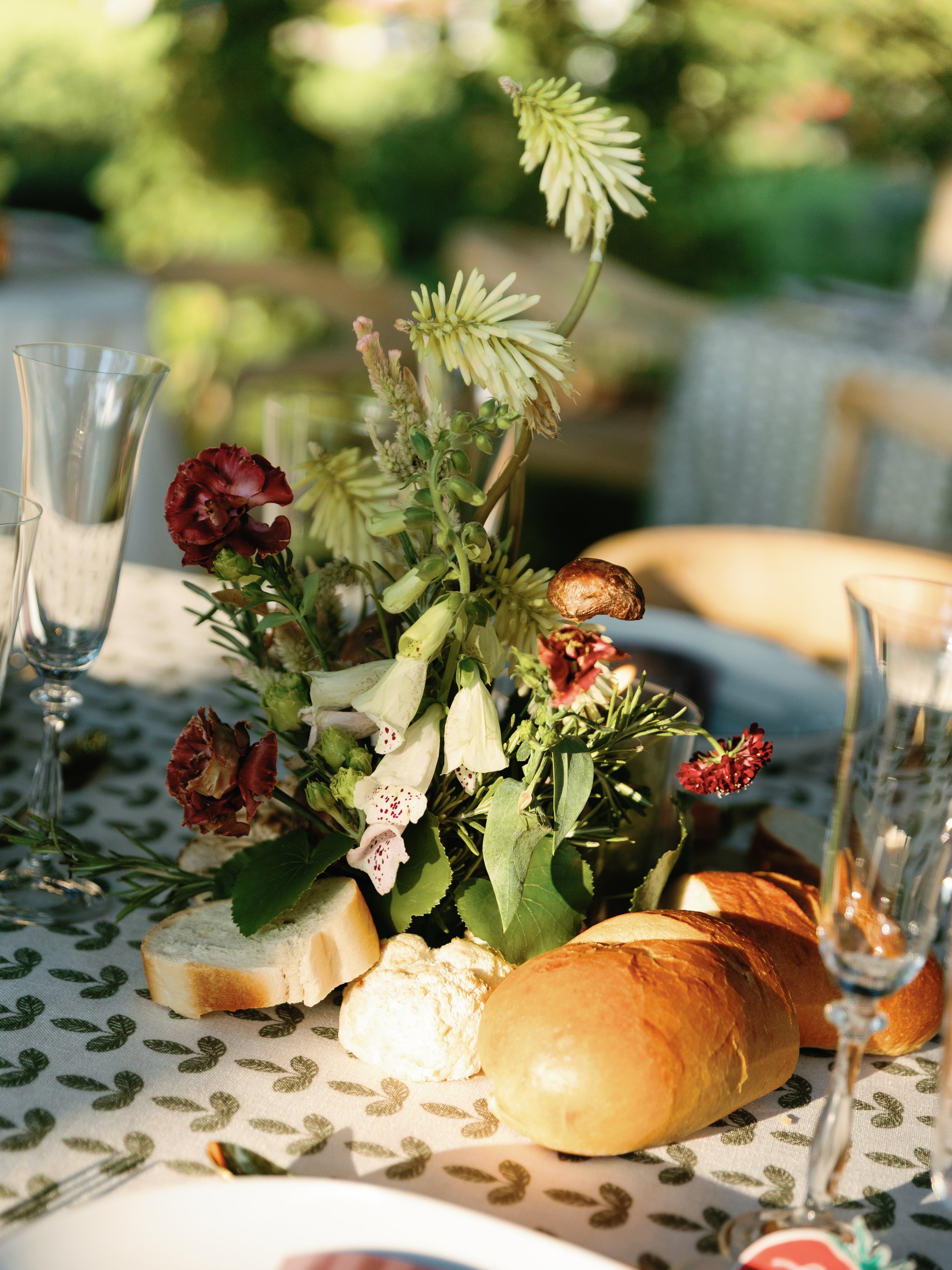 Elegant table setting with a floral centerpiece, bread rolls, and champagne glasses outdoors.