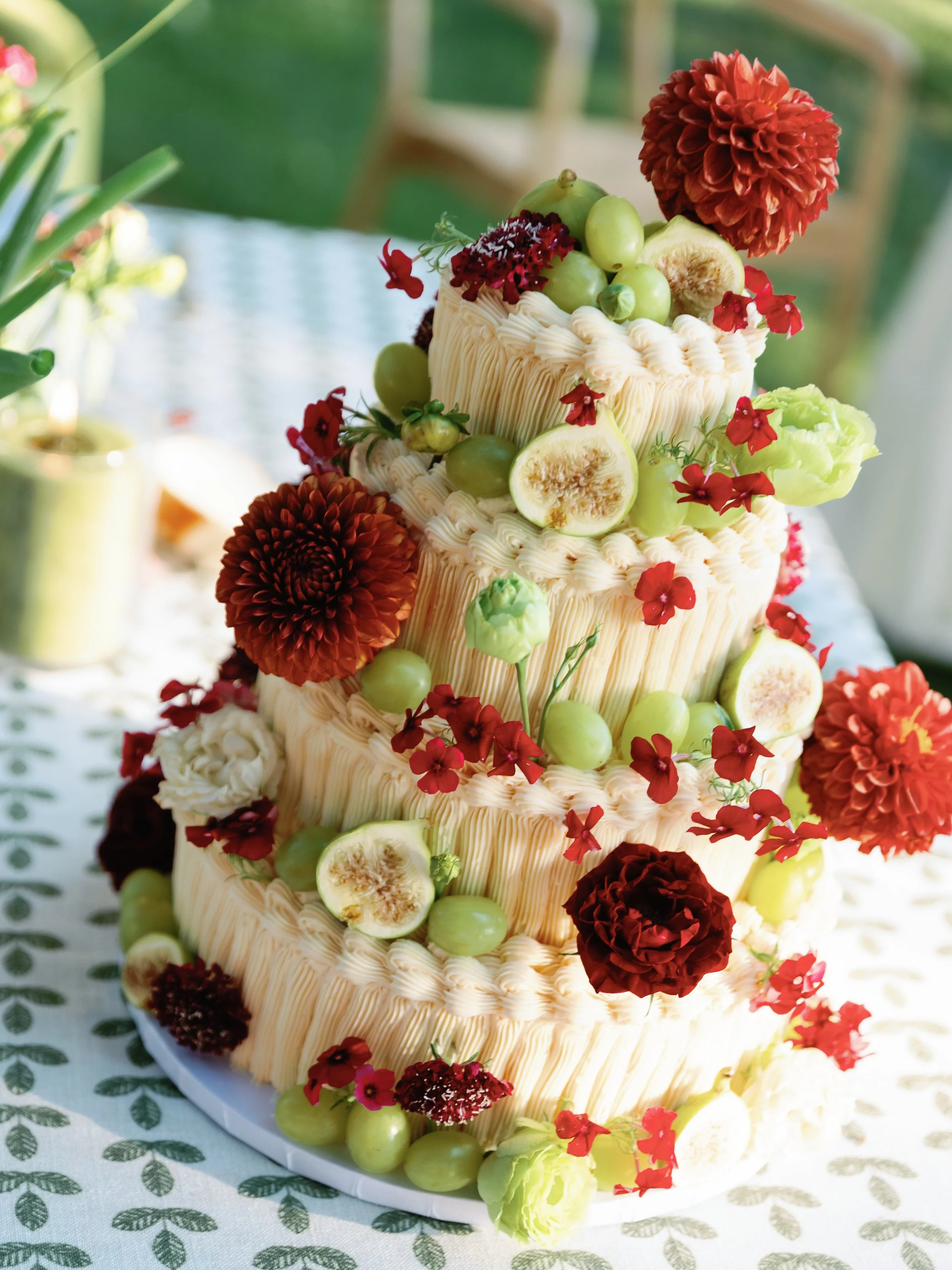 Three-tiered wedding cake decorated with figs, green grapes, and red flowers, sitting on a white cake stand.