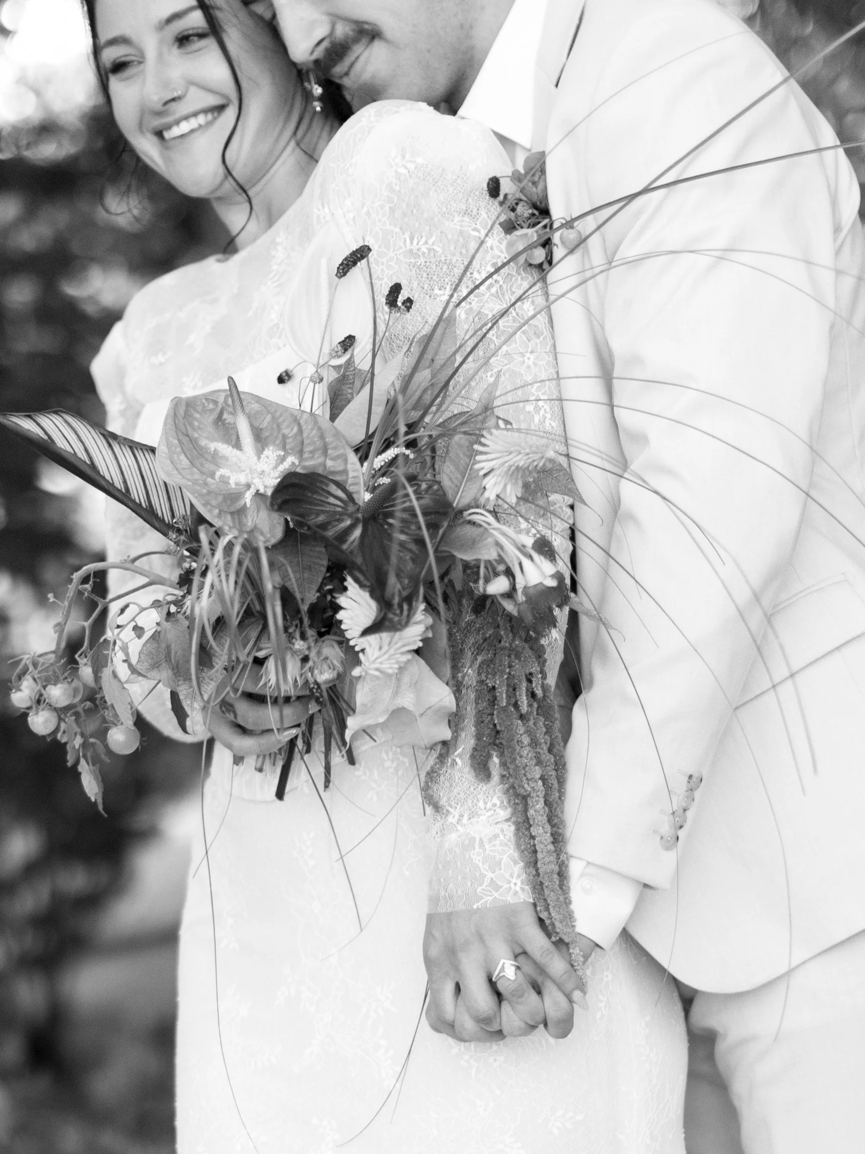Close-up of a bride and groom holding hands, showing the bride's wedding dress, bouquet, and engagement ring, smiling and embracing each other.