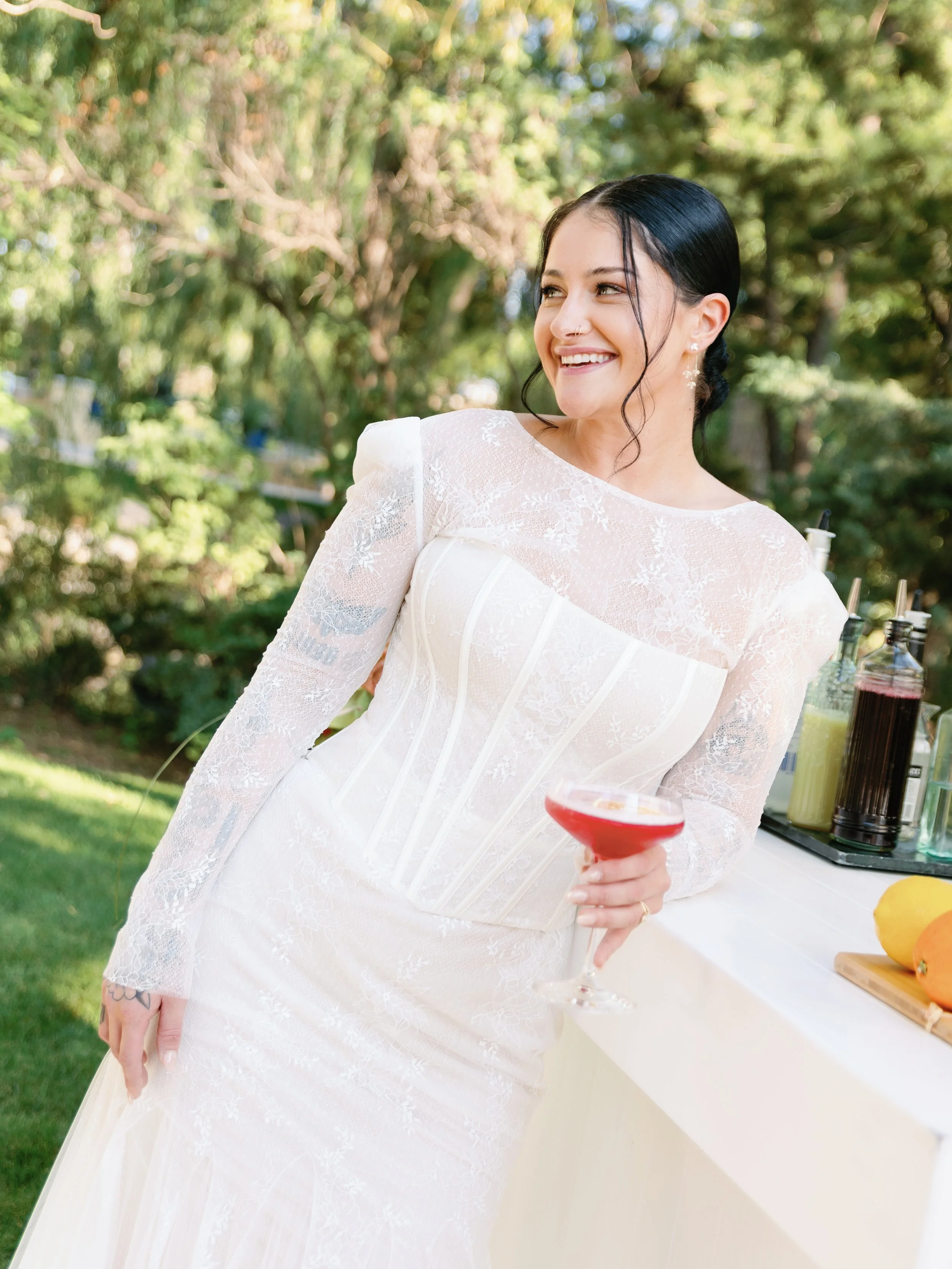A woman in a white lace wedding dress holding a pink cocktail, smiling outdoors with green trees in the background.