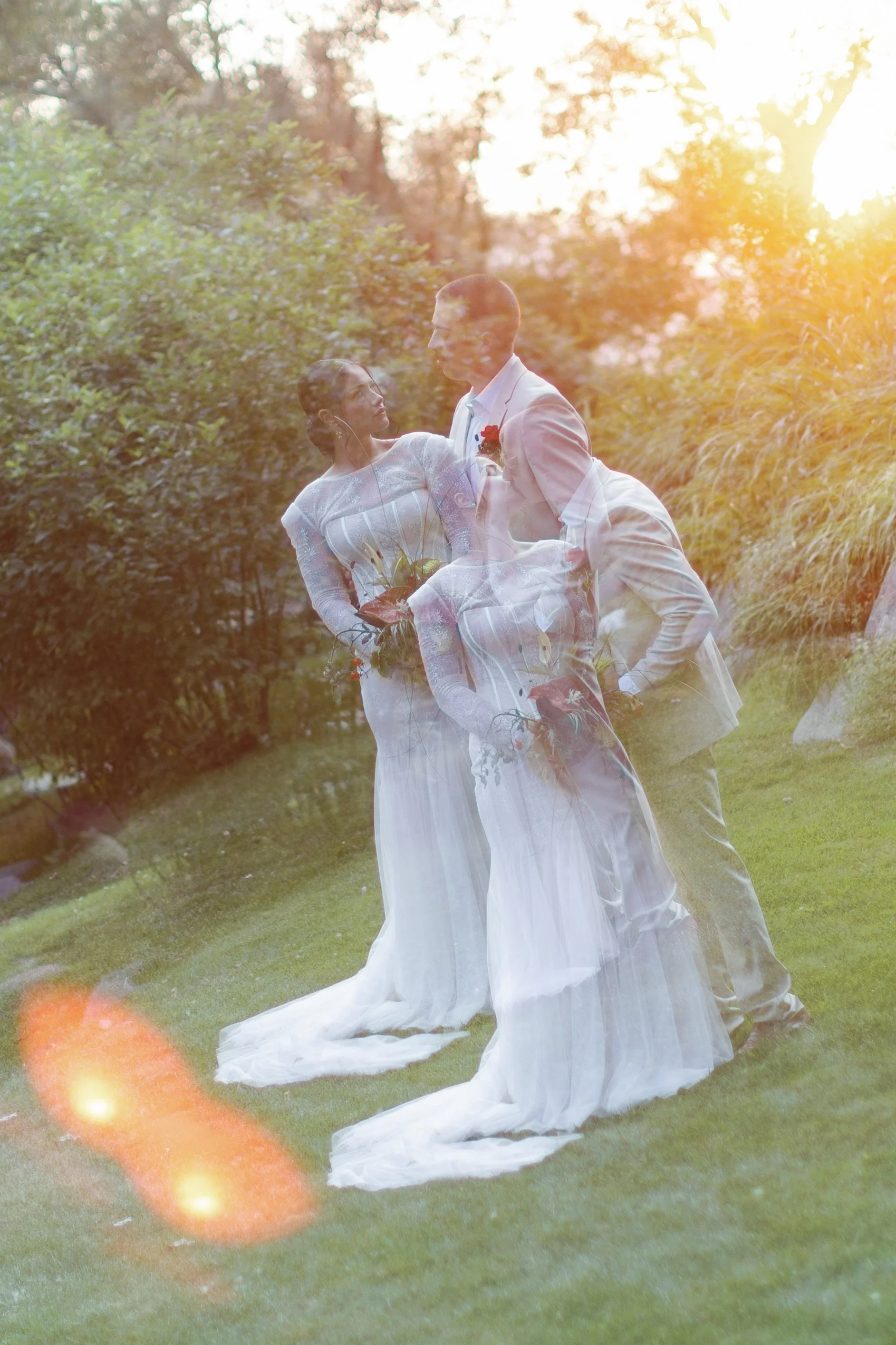 A couple in wedding attire, holding hands and gazing at each other, standing on grass with trees and bushes in the background, captured in a dreamlike, layered photo with sunlight flare.
