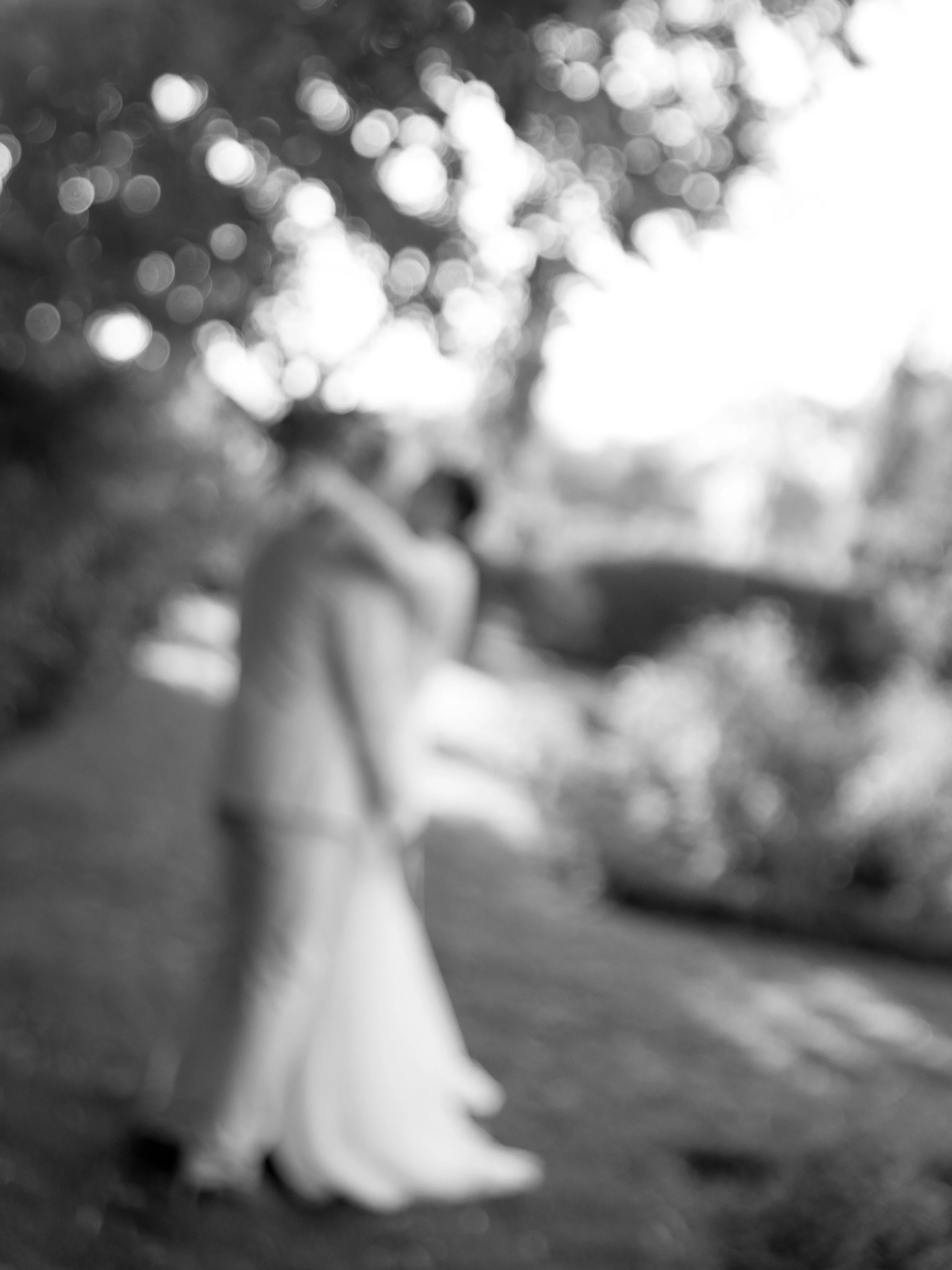 A blurry black and white photo of a couple standing close together outdoors, with trees and sunlight in the background.