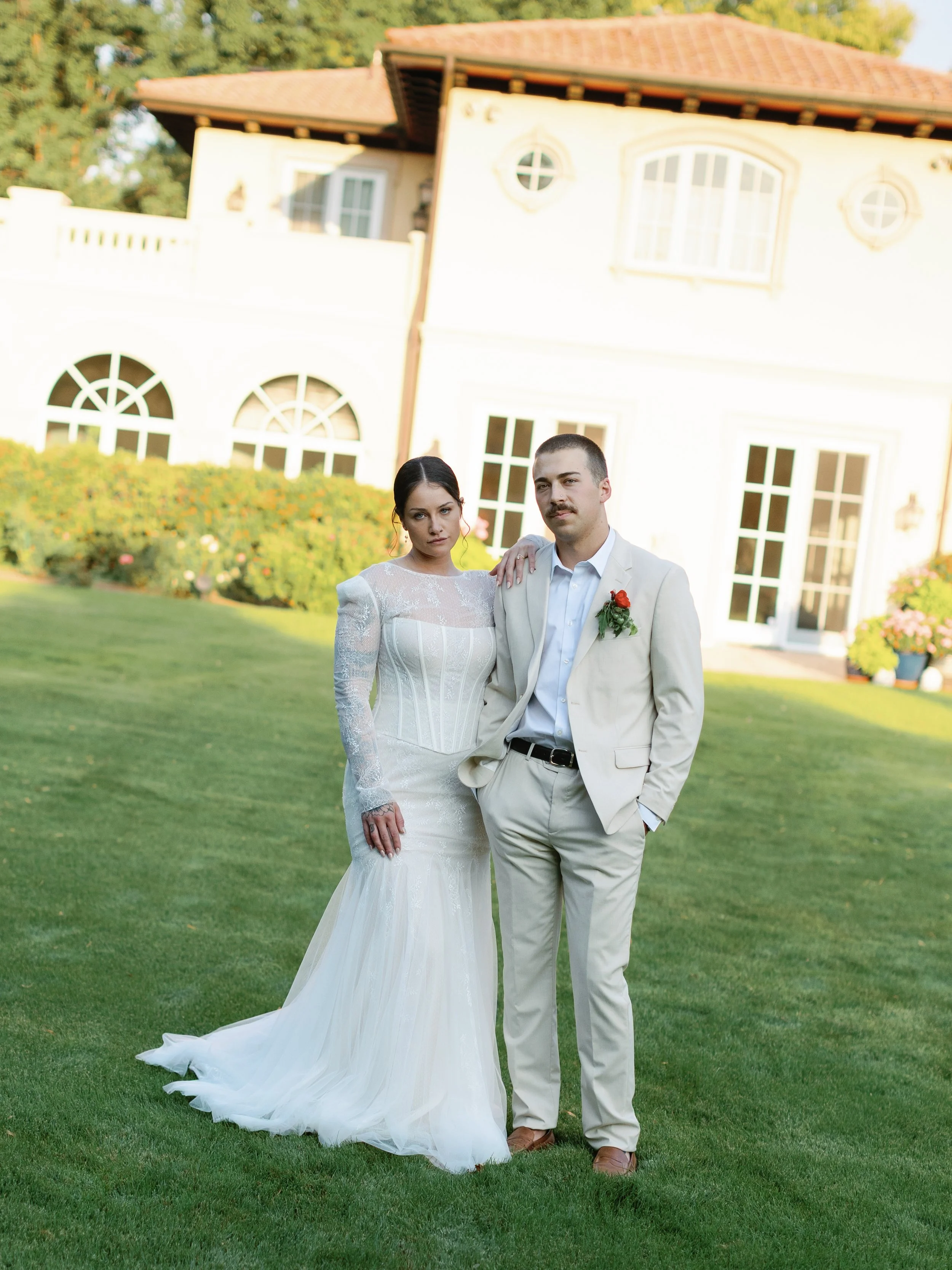 A bride and groom standing on a grassy lawn in front of a large house, with the bride wearing a white wedding dress and the groom in a cream-colored suit with a red boutonniere.