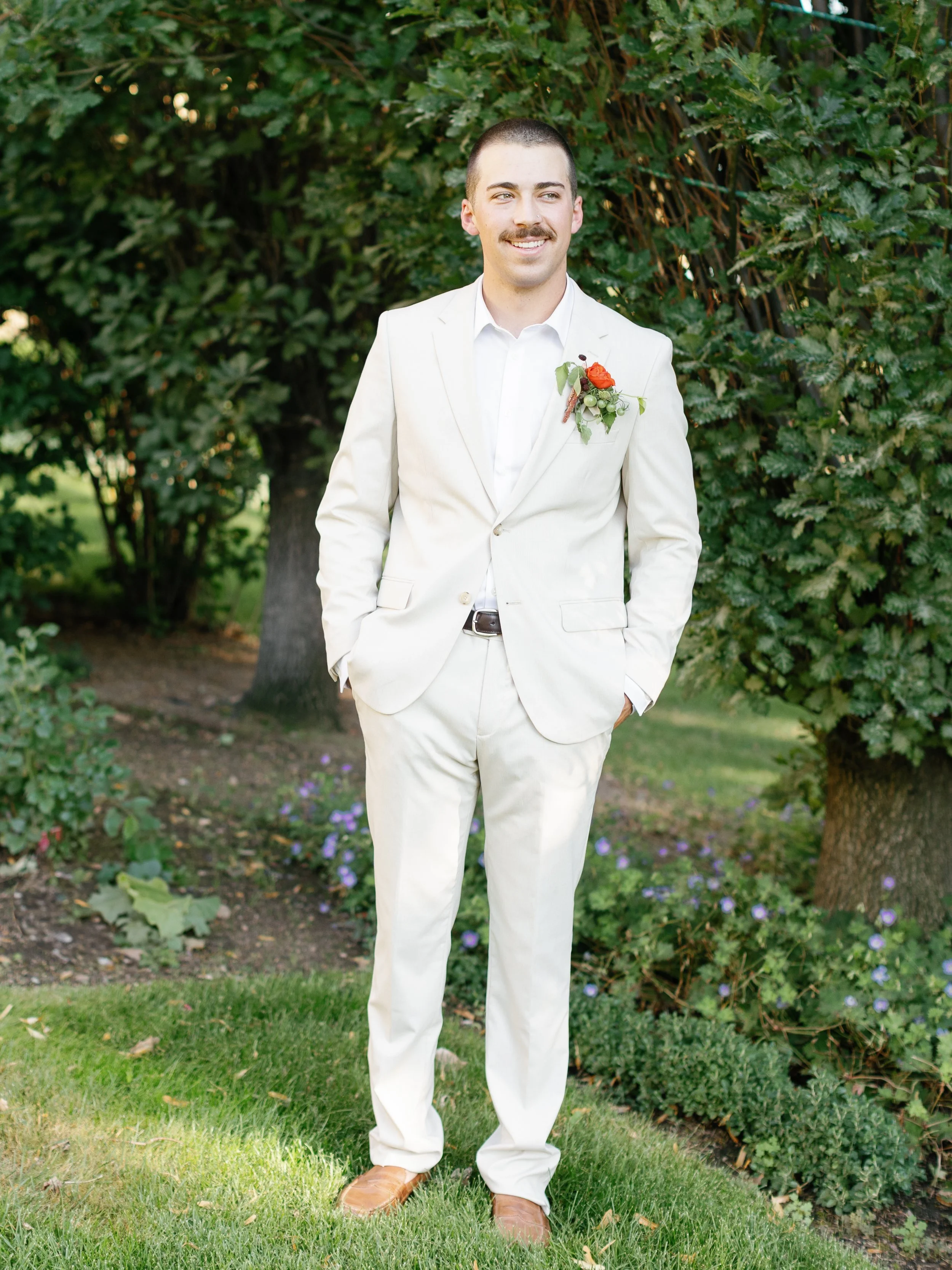 A man in a white suit with a boutonniere standing outdoors in a garden with trees and purple flowers.