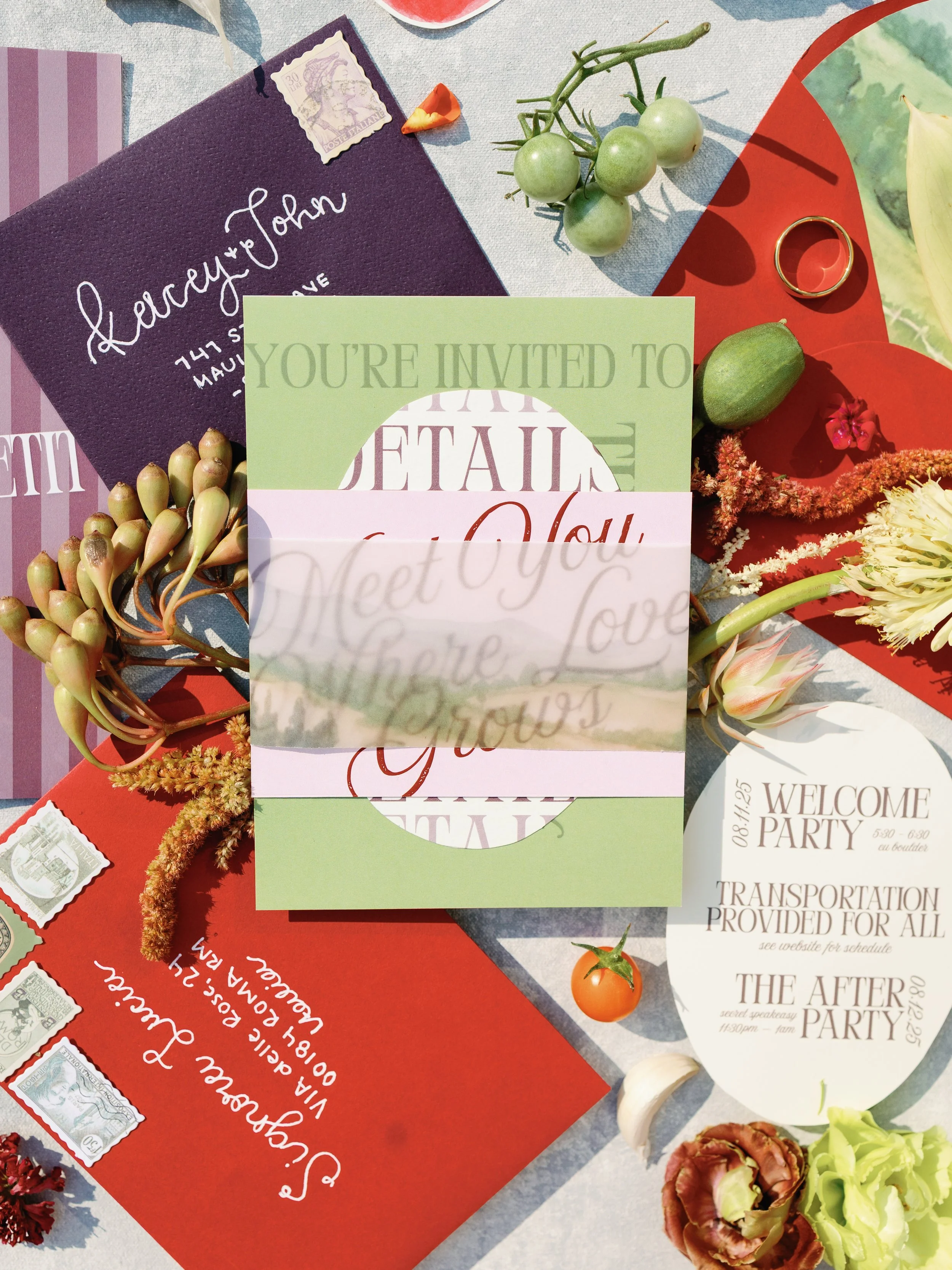 Wedding invitation suite with an RSVP card, welcome party invitation, and details card surrounded by flowers, a ring, and grapes on a light-colored surface.