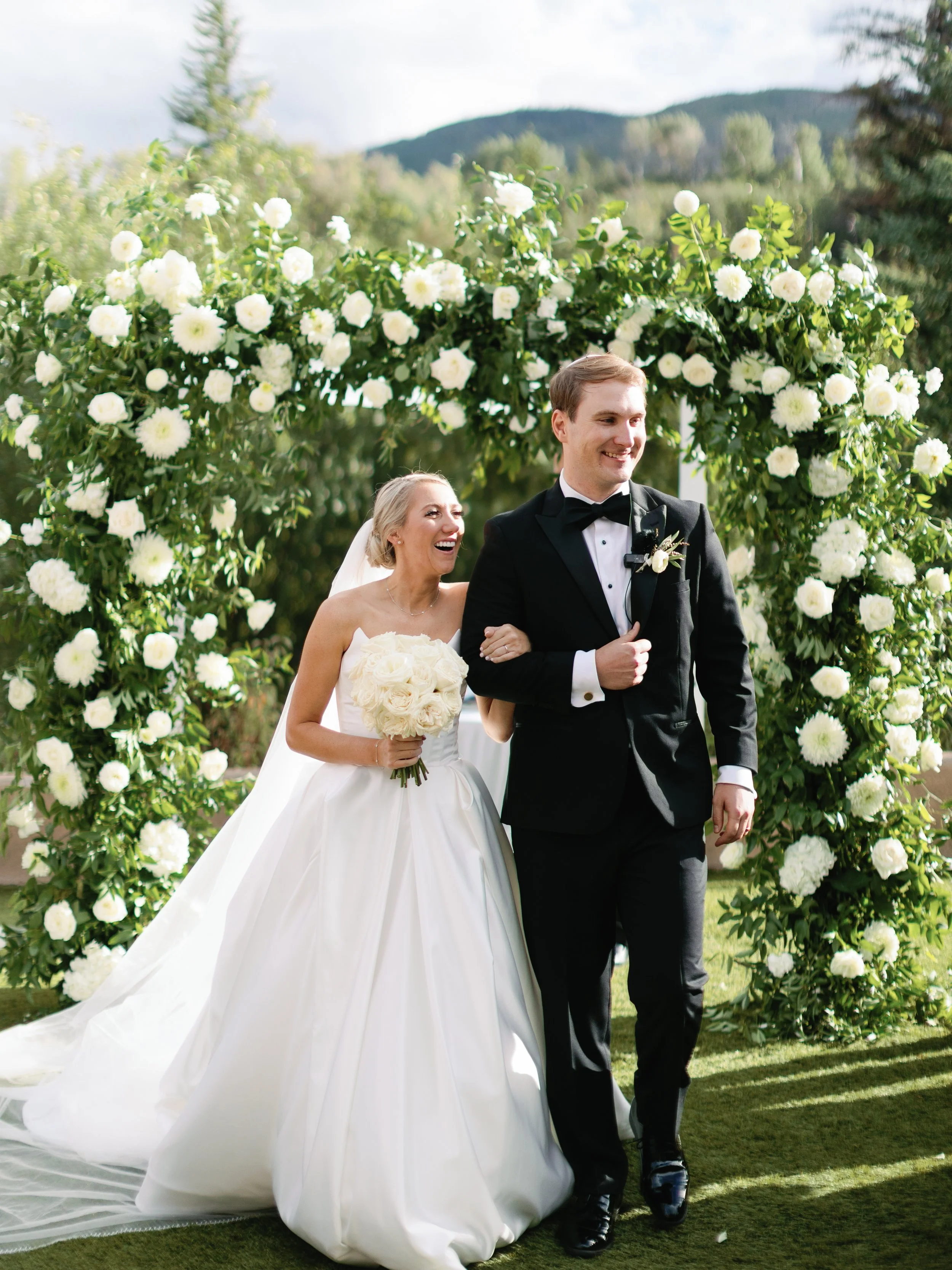 A bride and groom at their outdoor wedding ceremony, with the bride holding a bouquet of white flowers and the groom walking arm-in-arm with her through a floral archway of white roses and greenery, against a backdrop of mountains and trees.