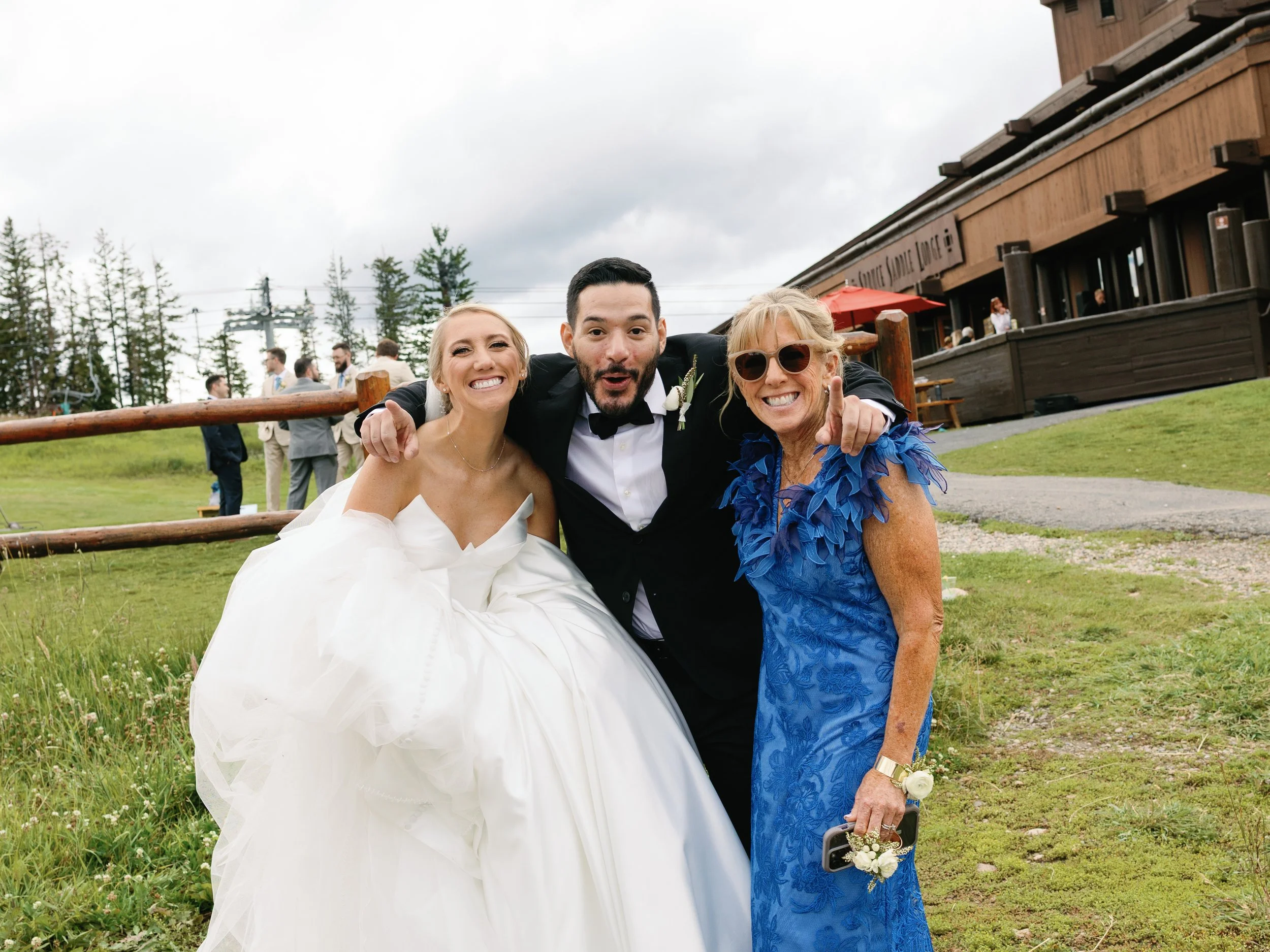 Three people smiling and posing outdoors at a wedding, with the bride in a white wedding gown, the groom in a black tuxedo, and a woman in a blue dress with lace detailing and sunglasses.