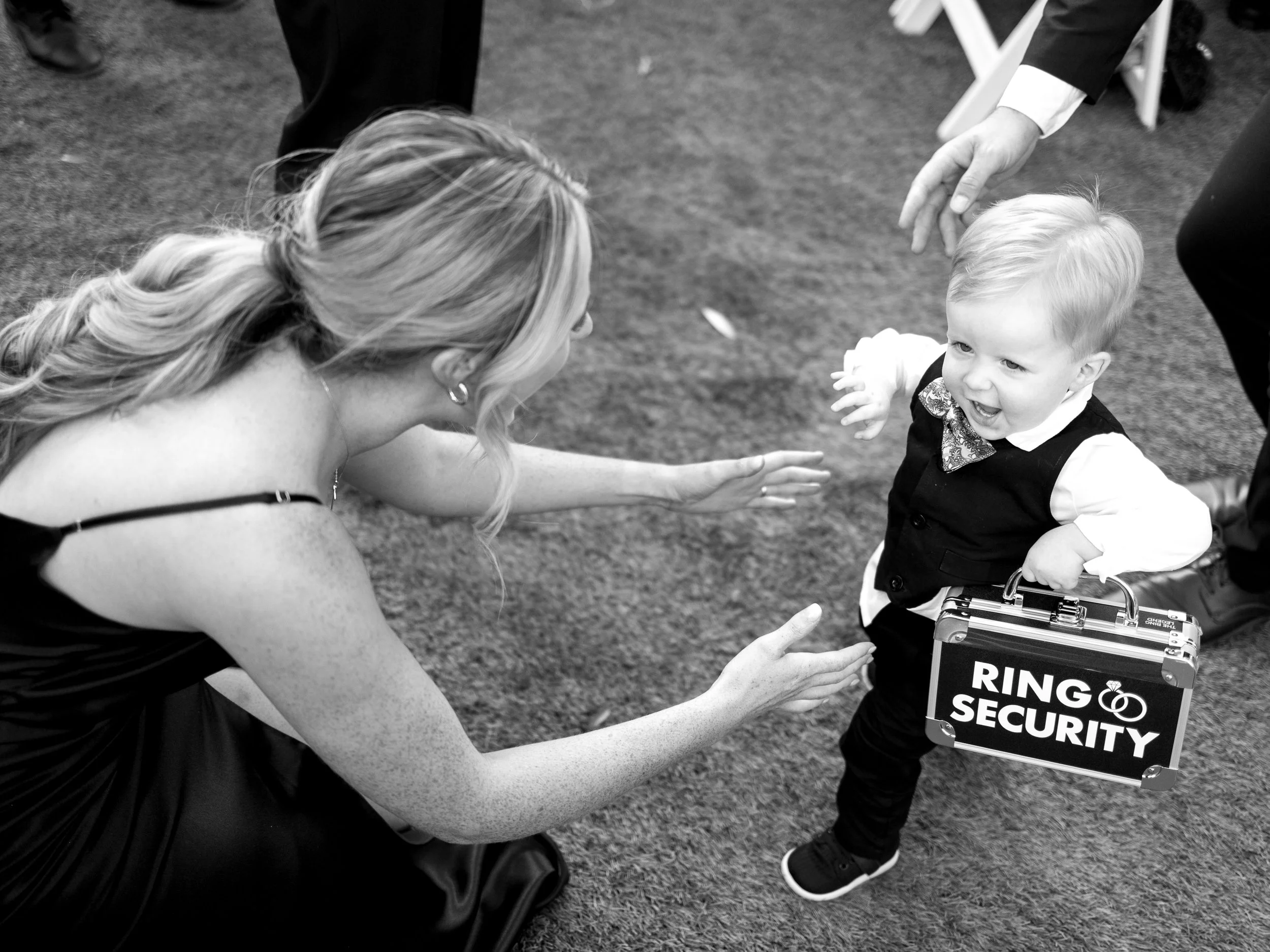 A woman in a black dress is crouched down, reaching out to a young boy dressed in formal attire with a "Ring Security" briefcase, who is smiling and holding out his hand in a playful manner.