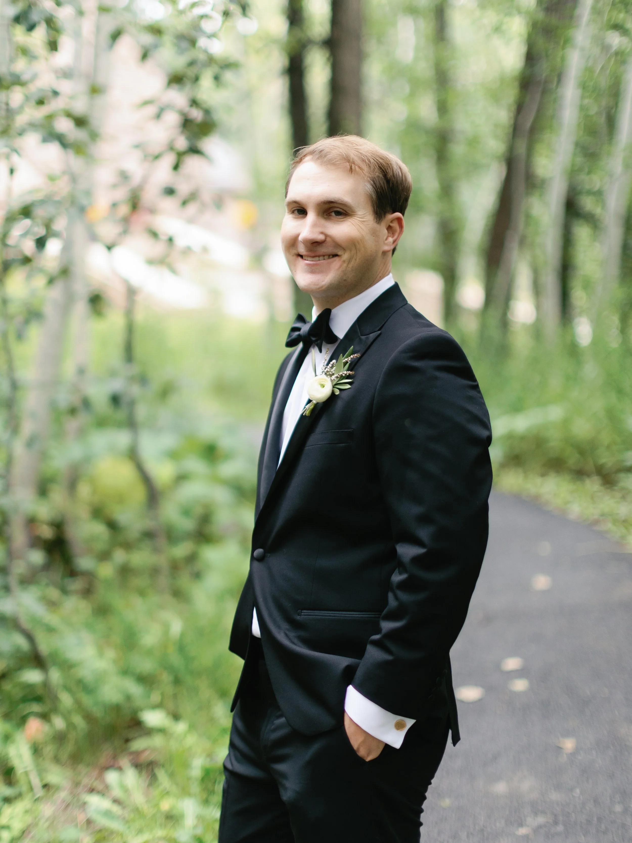 A man in a black tuxedo, white shirt, and black bow tie, standing on a wooded pathway with greenery, smiling and looking at the camera.