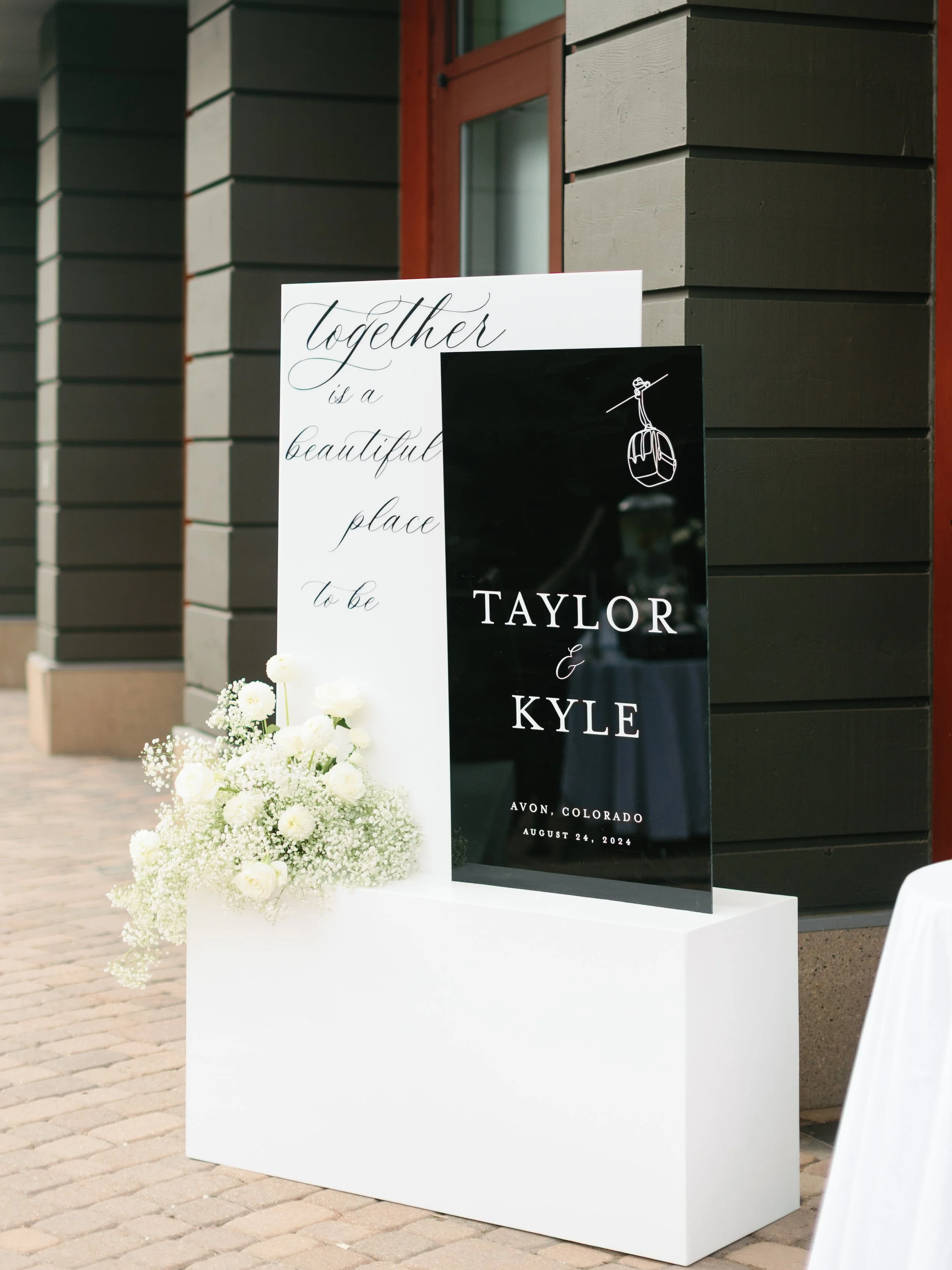 Wedding sign with white flowers on a white stand, featuring black and white text for Taylor and Kyle's wedding on August 24, 2024, in Avon, Colorado, outside a building with dark gray siding.