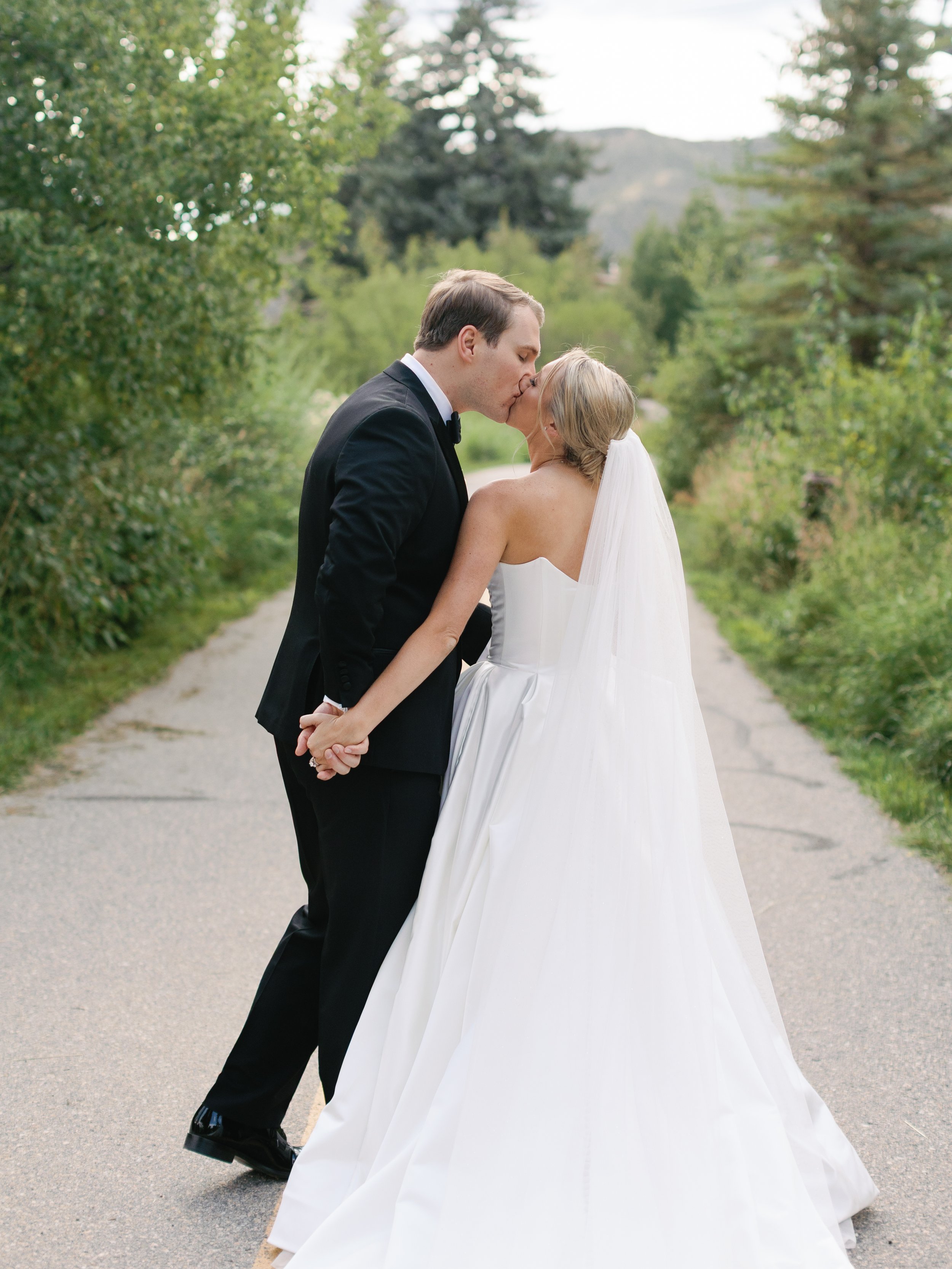 A newlywed couple, dressed in formal wedding attire, sharing a kiss and holding hands on a rural road with greenery and trees.
