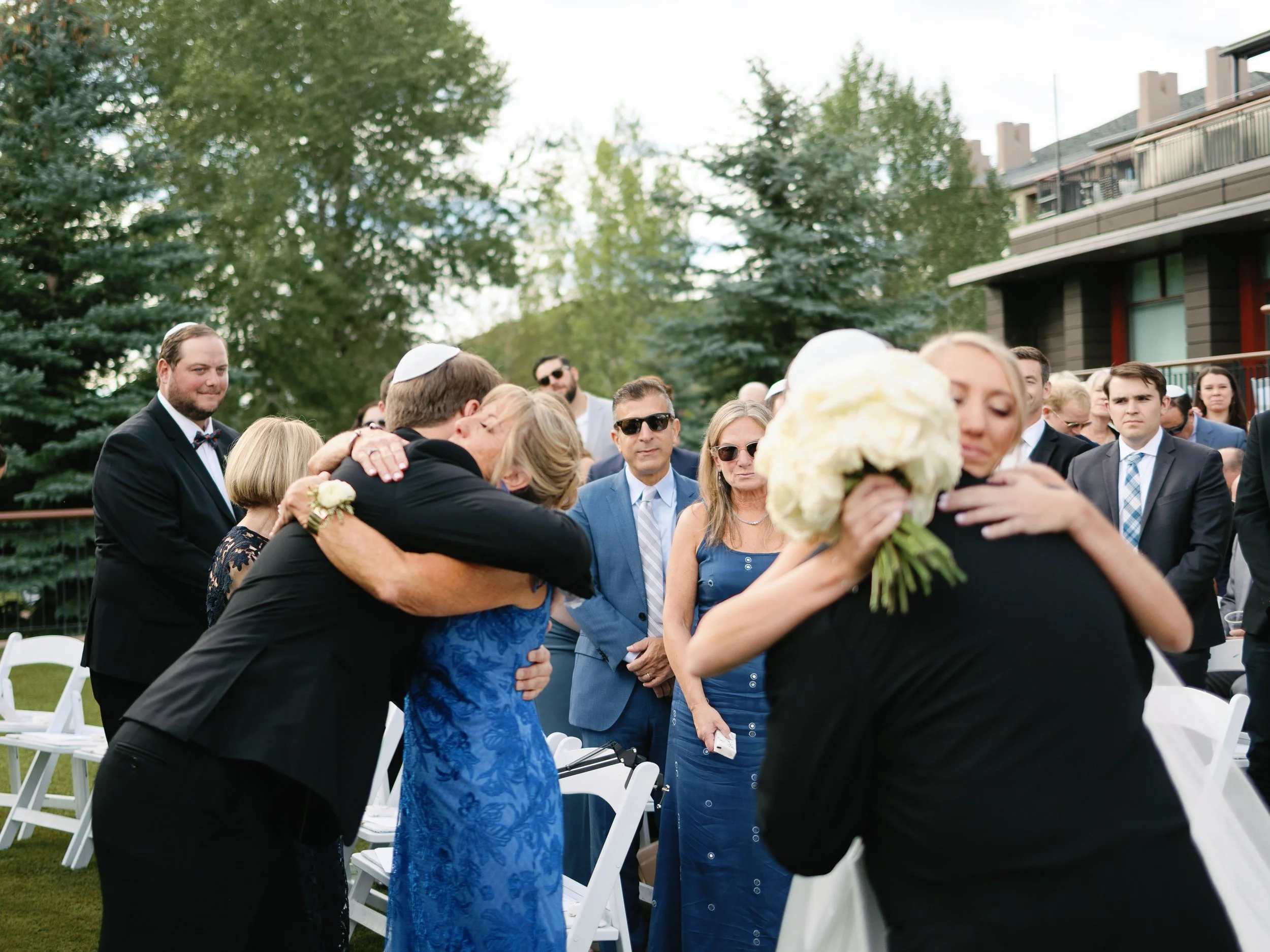 People at a wedding outdoor ceremony emotionally hugging and embracing, with some guests watching in the background, trees, and a building.