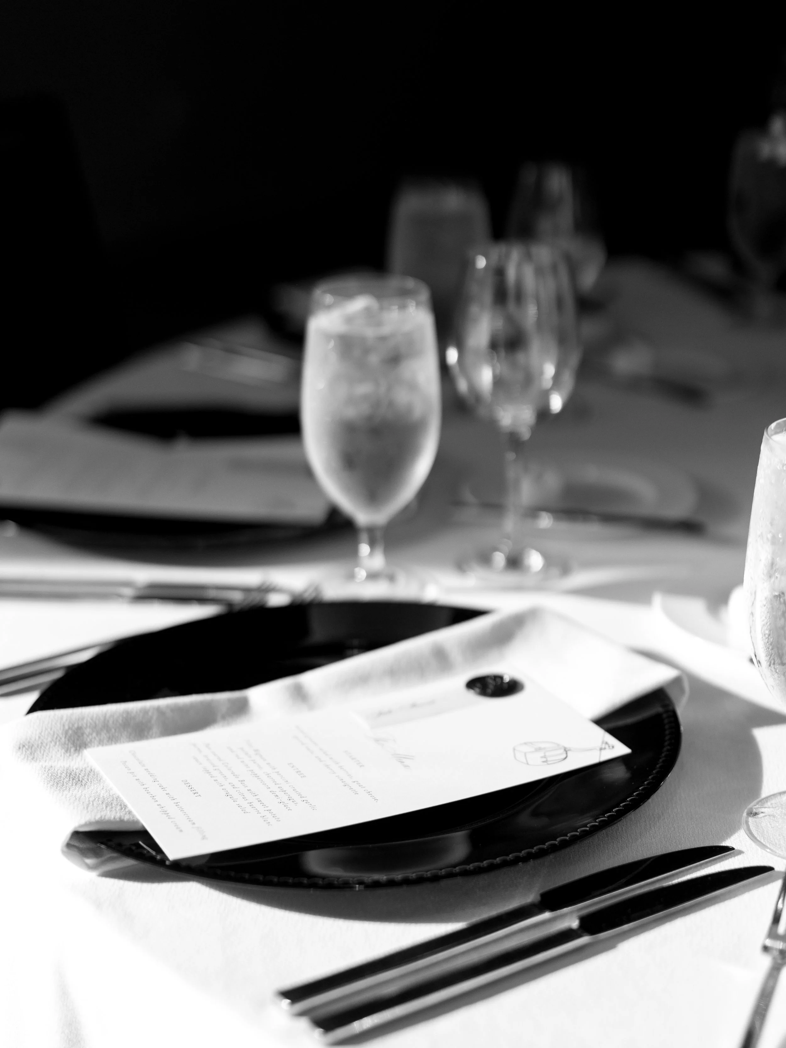 Elegant table setting with a black plate, a menu, silverware, and three glasses filled with water, on a white tablecloth.