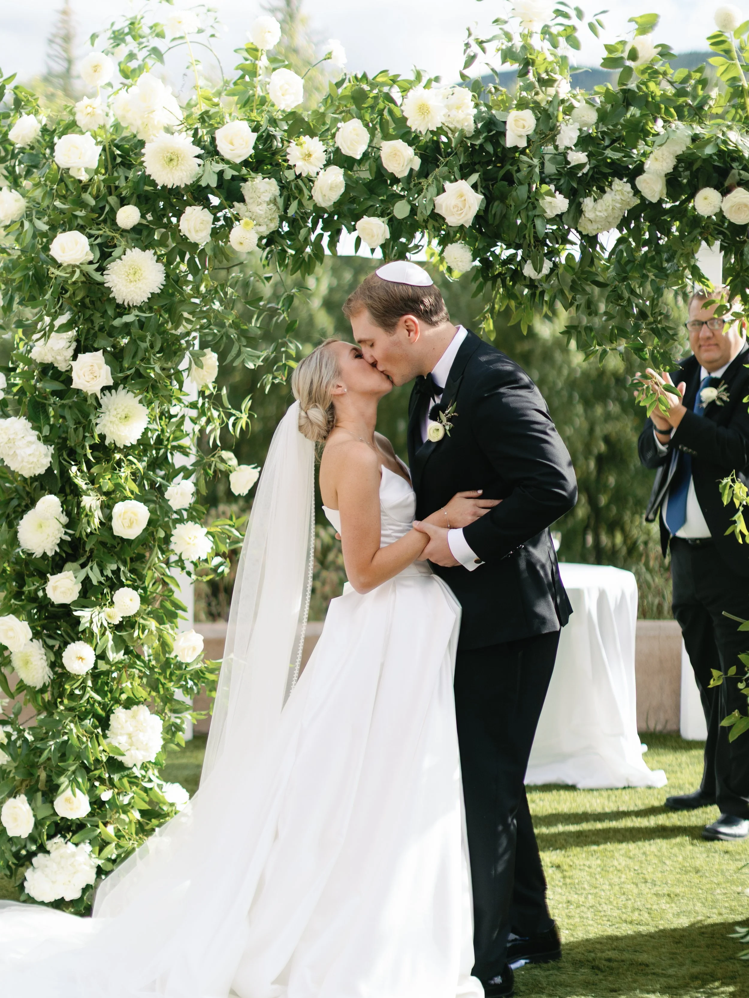 A couple is kissing during their outdoor wedding ceremony beneath a floral arch of white roses and greenery. The bride is wearing a white wedding gown and veil, and the groom is in a black tuxedo. An officiant stands nearby, clapping.