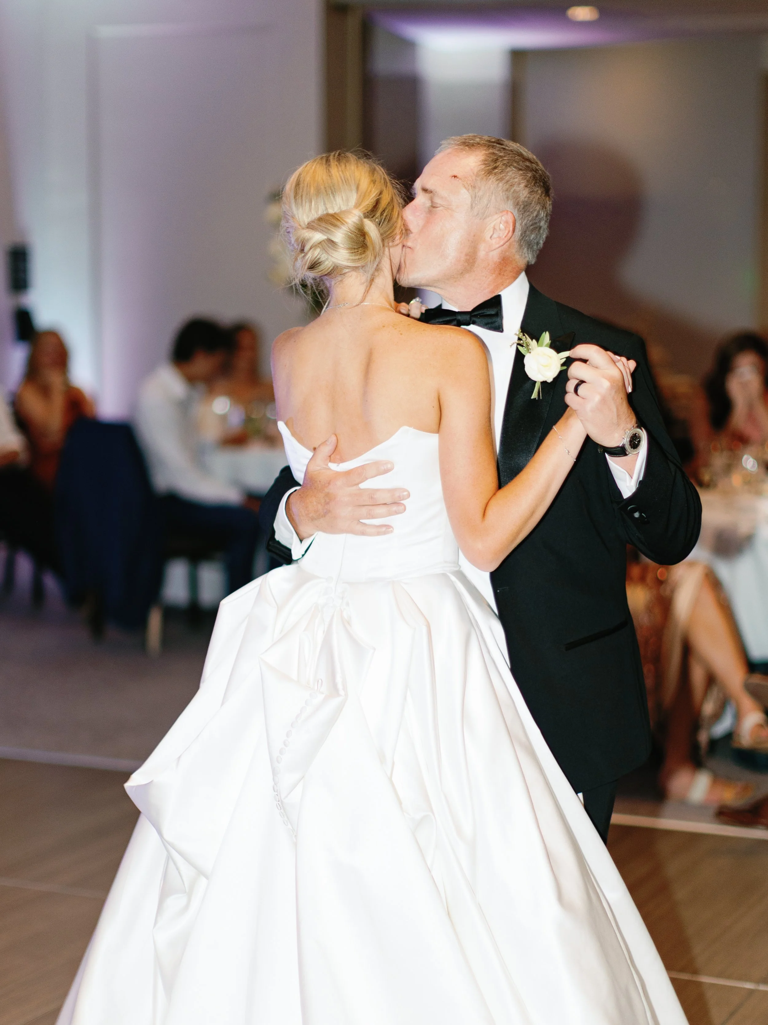 A bride and groom sharing a kiss during their wedding dance surrounded by guests.