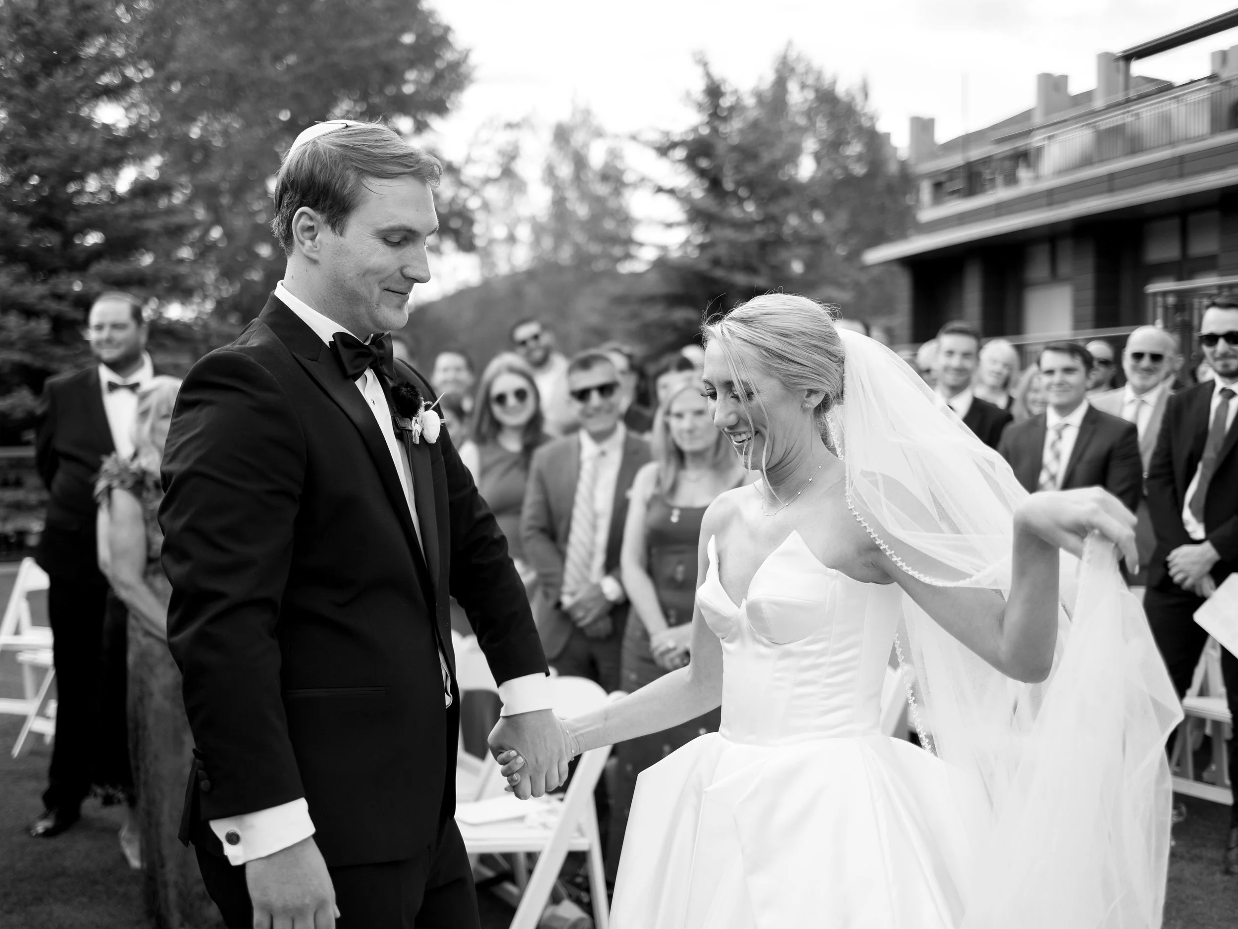 A black-and-white photo of a bride and groom holding hands during their wedding ceremony outdoors, with guests in the background.