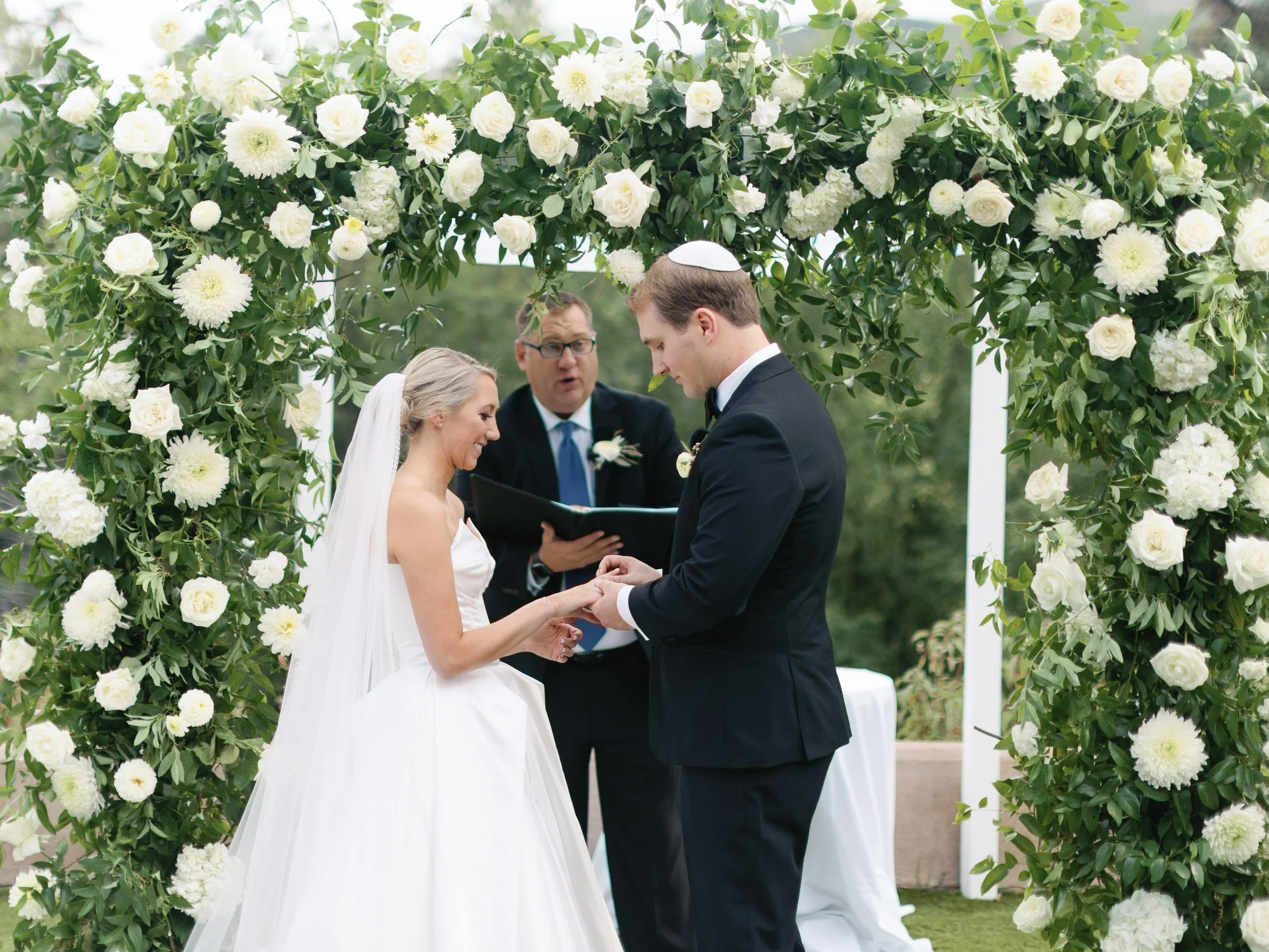 A couple exchanging wedding vows under a lush floral arch at an outdoor wedding ceremony with an officiant in the background.