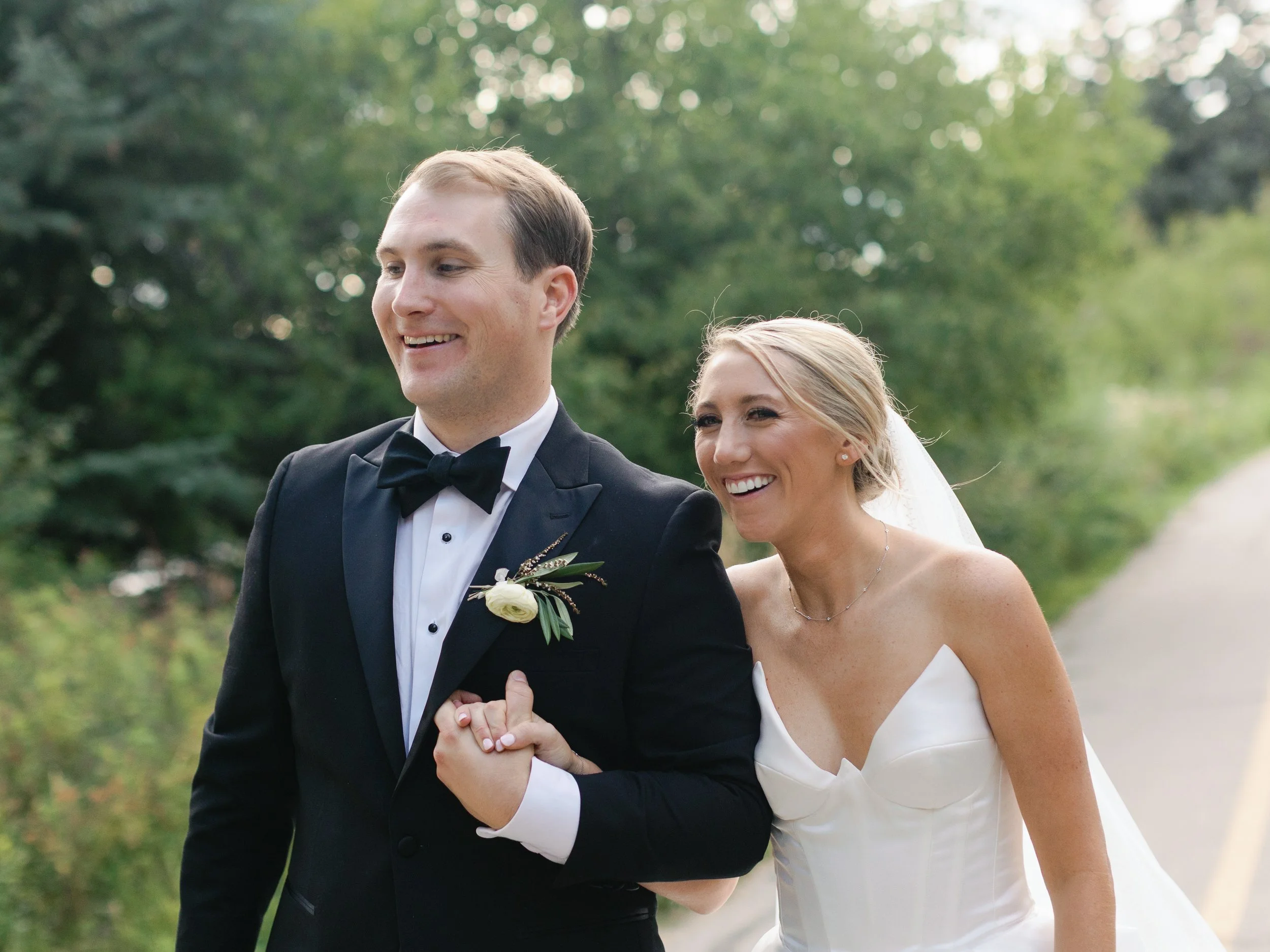 A smiling bride and groom holding hands outdoors during their wedding, with green trees in the background.