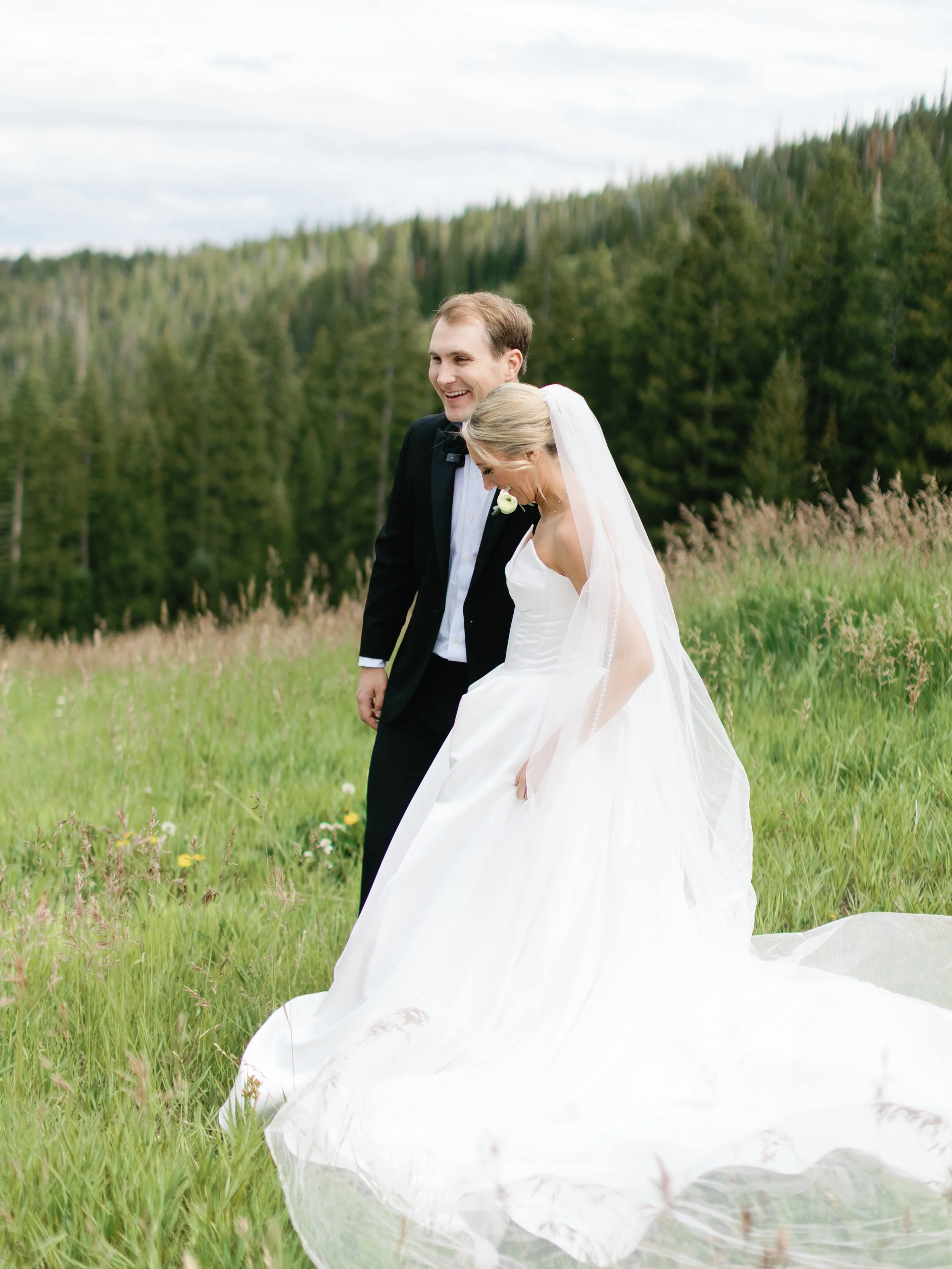 A bride and groom walking in a grassy field with wildflowers and a forested mountain in the background.