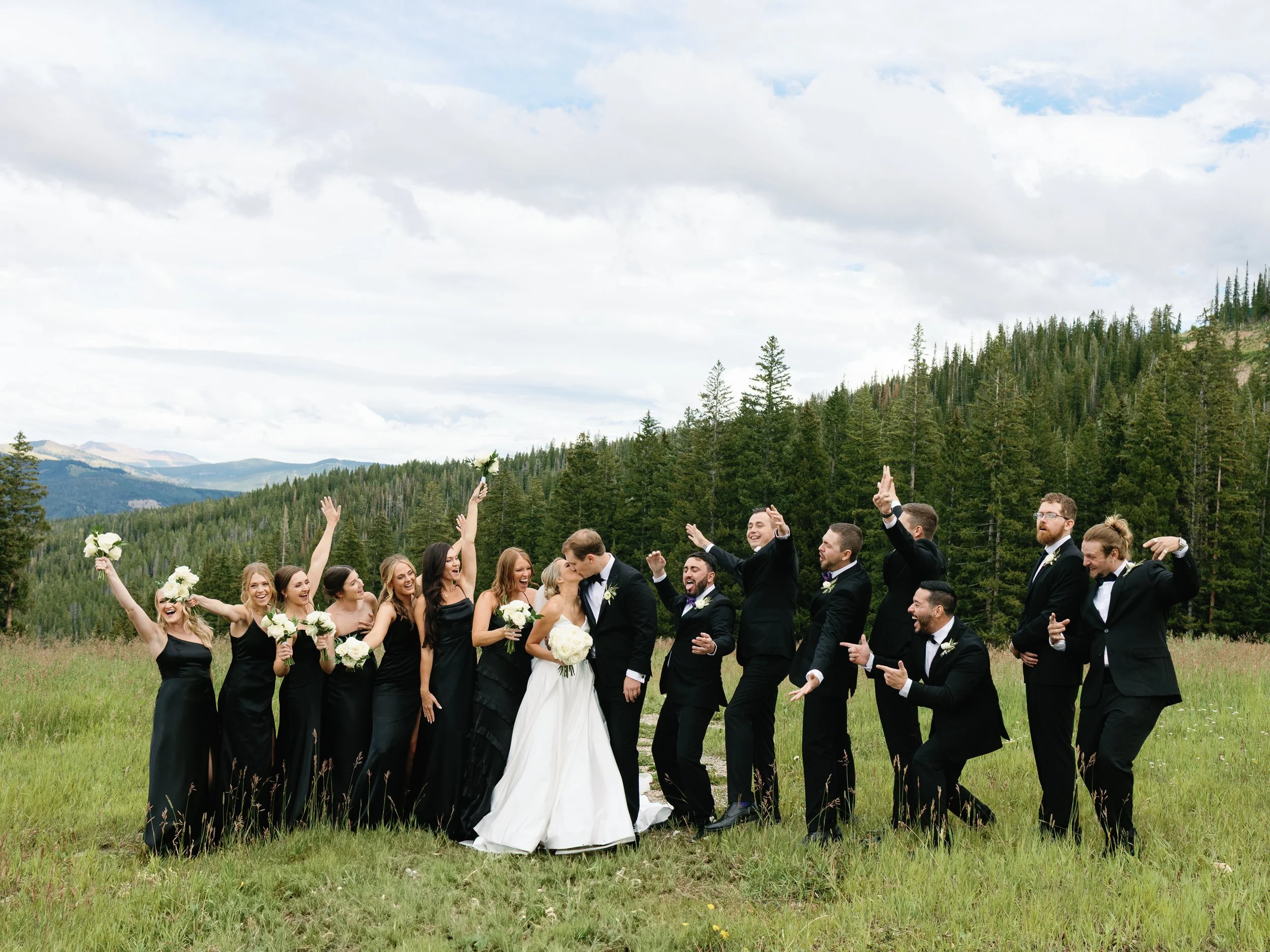 A group of wedding guests, including women in black dresses and men in black suits, celebrating outdoors on a grassy field with a mountain and forest background. The bride and groom are in the center, kissing, while the guests cheer and pose around t