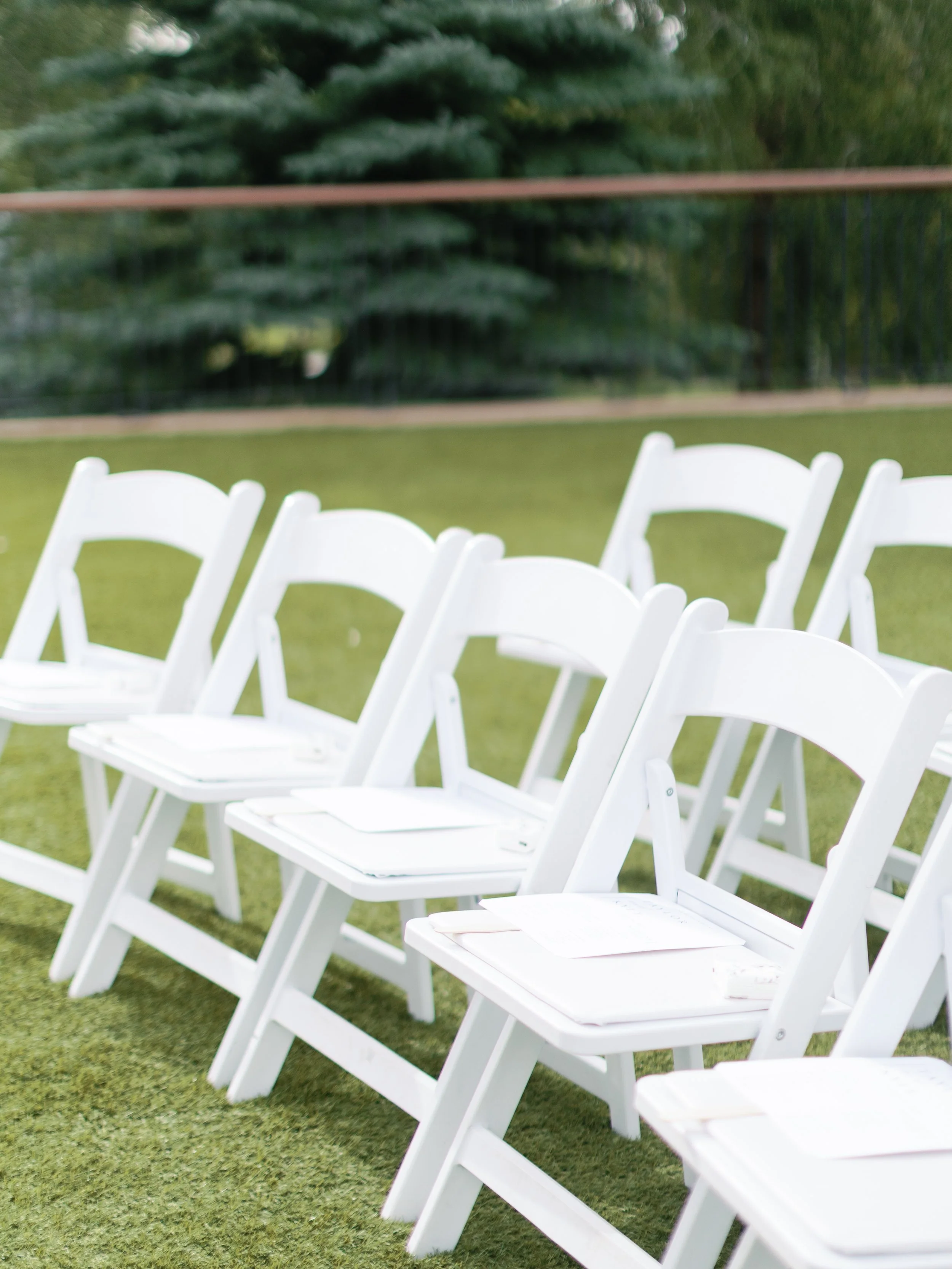 White folding chairs arranged in rows on a grassy area, with some papers on their seats, outdoors with a tree and wooden fence in the background.