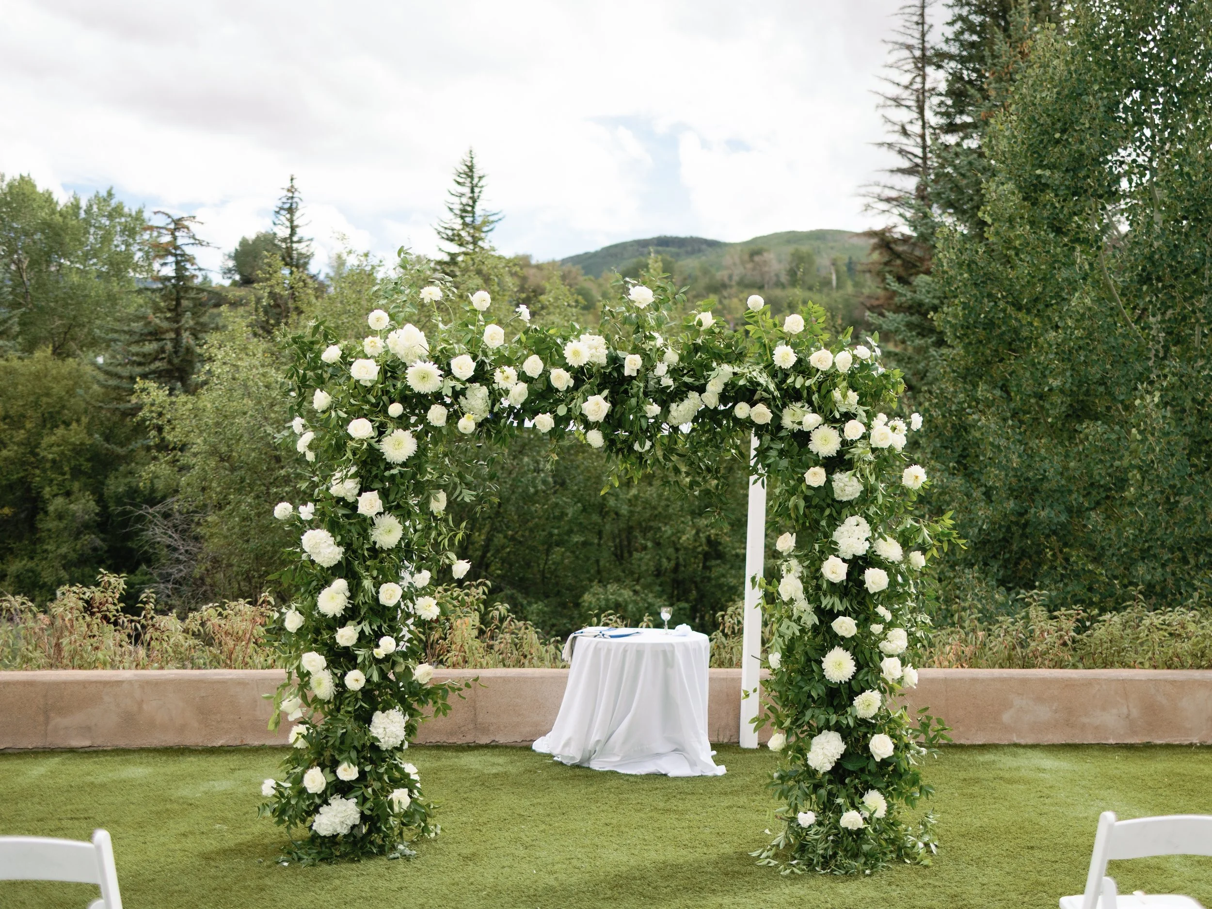 A wedding arch decorated with white flowers outdoors, with a small round table covered with a white cloth underneath, and greenery in the background.