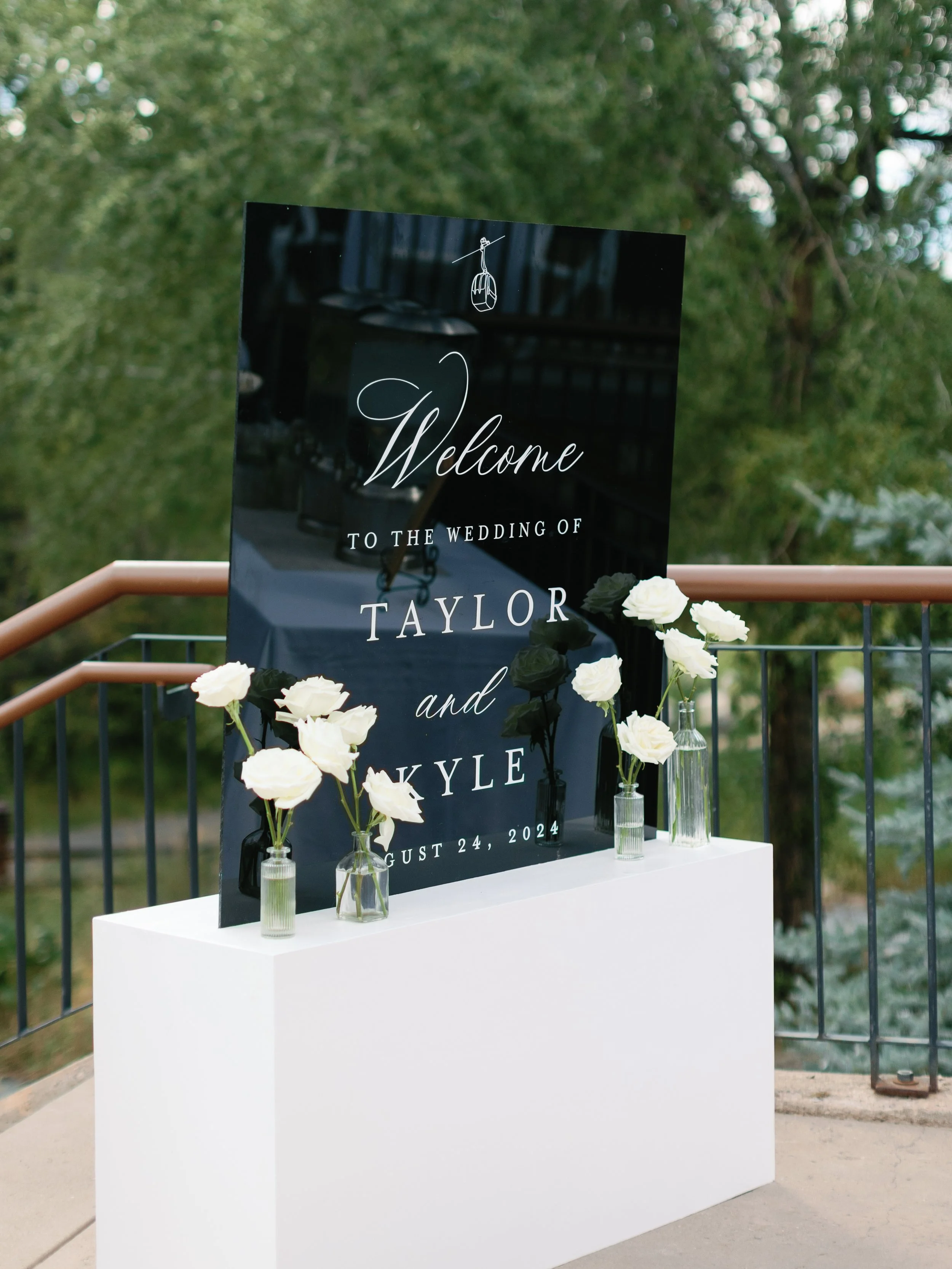 A wedding welcome sign displayed on a black reflective board with white floral arrangements in glass vases, reading "Welcome to the wedding of Taylor and Kyle, August 24, 2024". The sign is situated outdoors on a white pedestal with a background of t