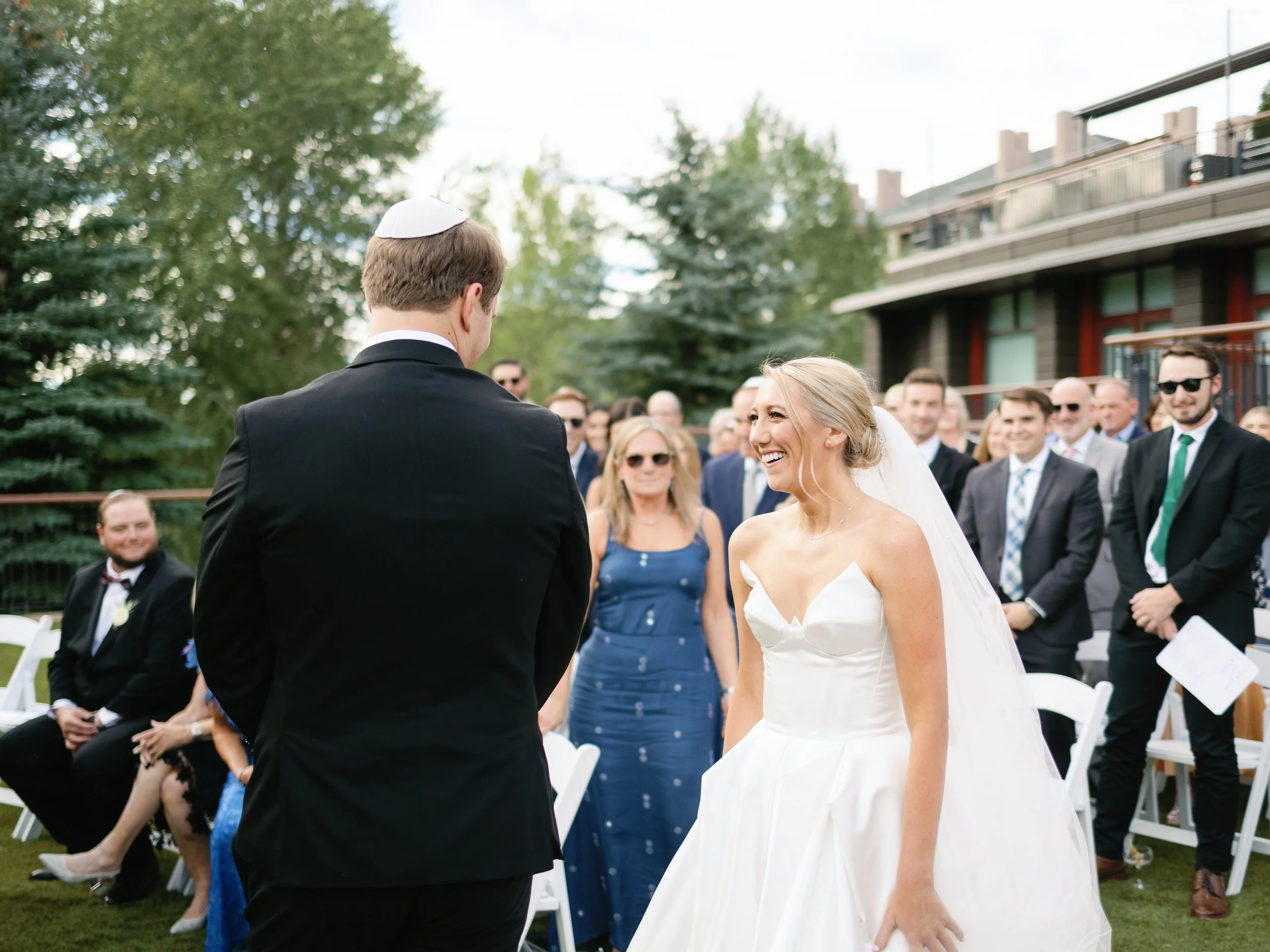 A bride and groom during an outdoor wedding ceremony. The bride is smiling and wearing a white dress and veil. The groom is dressed in a black suit and white kippah. Guests are seated and standing, watching the couple.