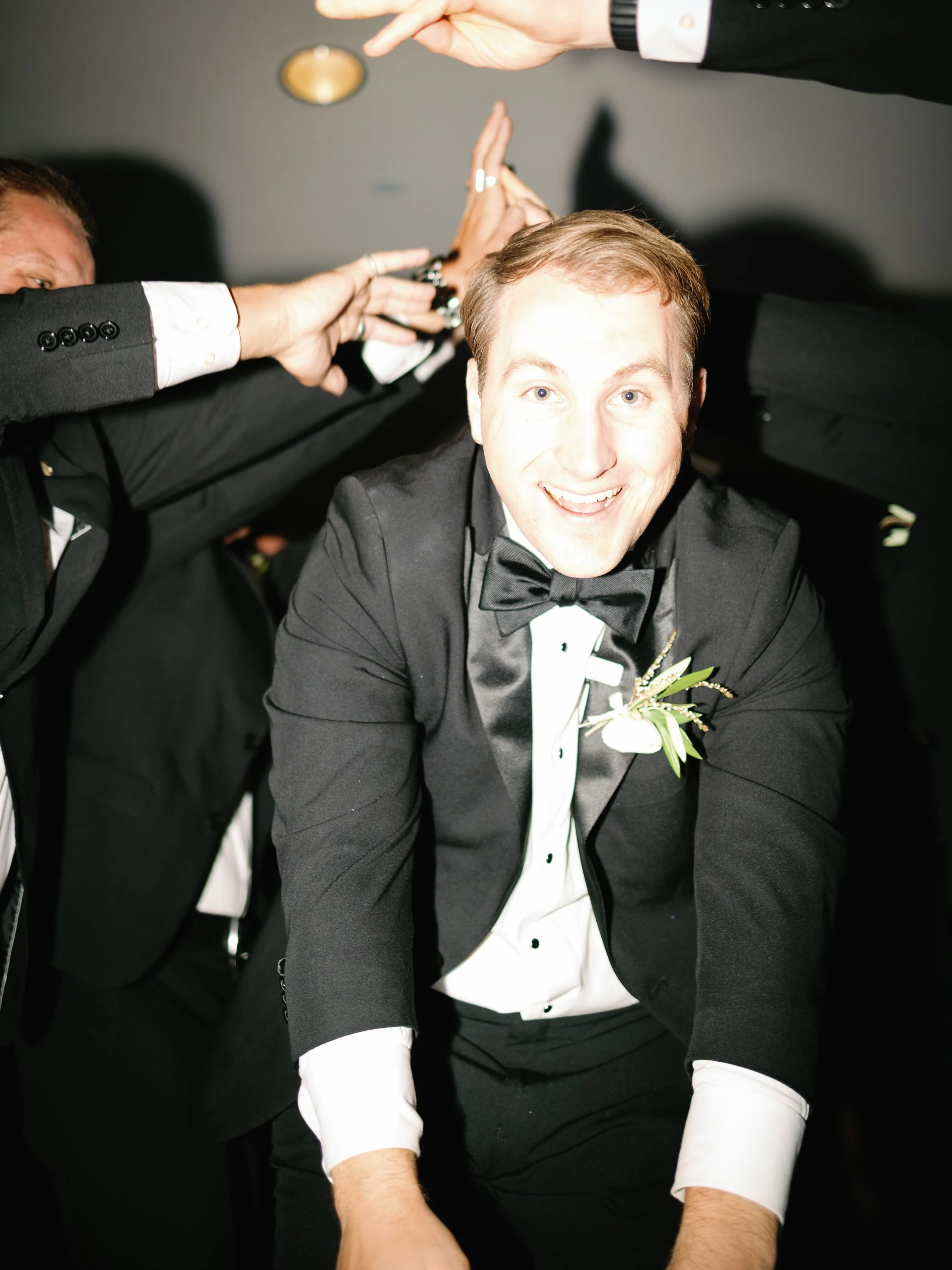 A man in a tuxedo with a bowtie and boutonniere is smiling while surrounded by people giving him high-fives.