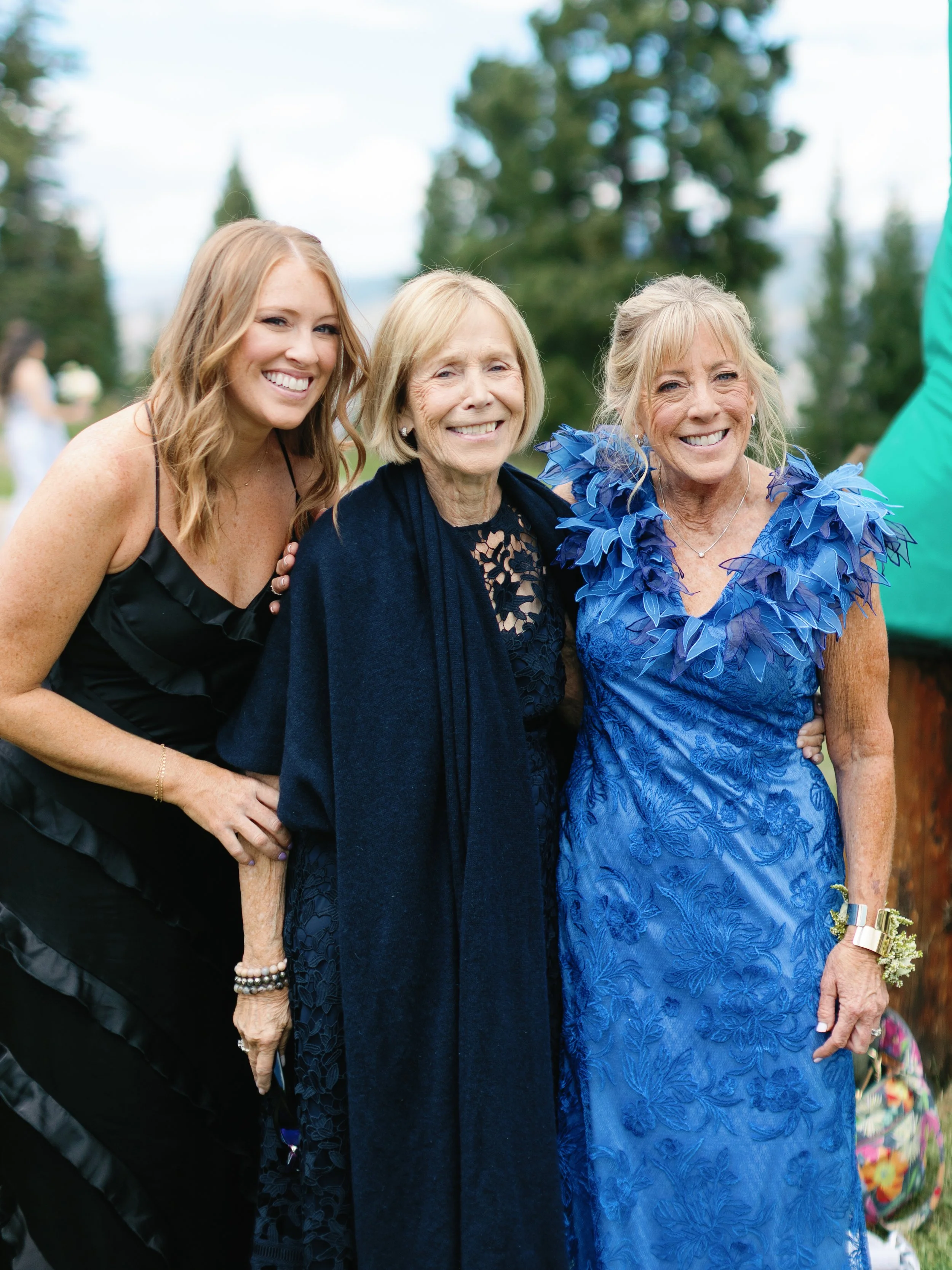 Three women smiling outdoors, dressed in formal attire, with trees and a blue sky in the background.