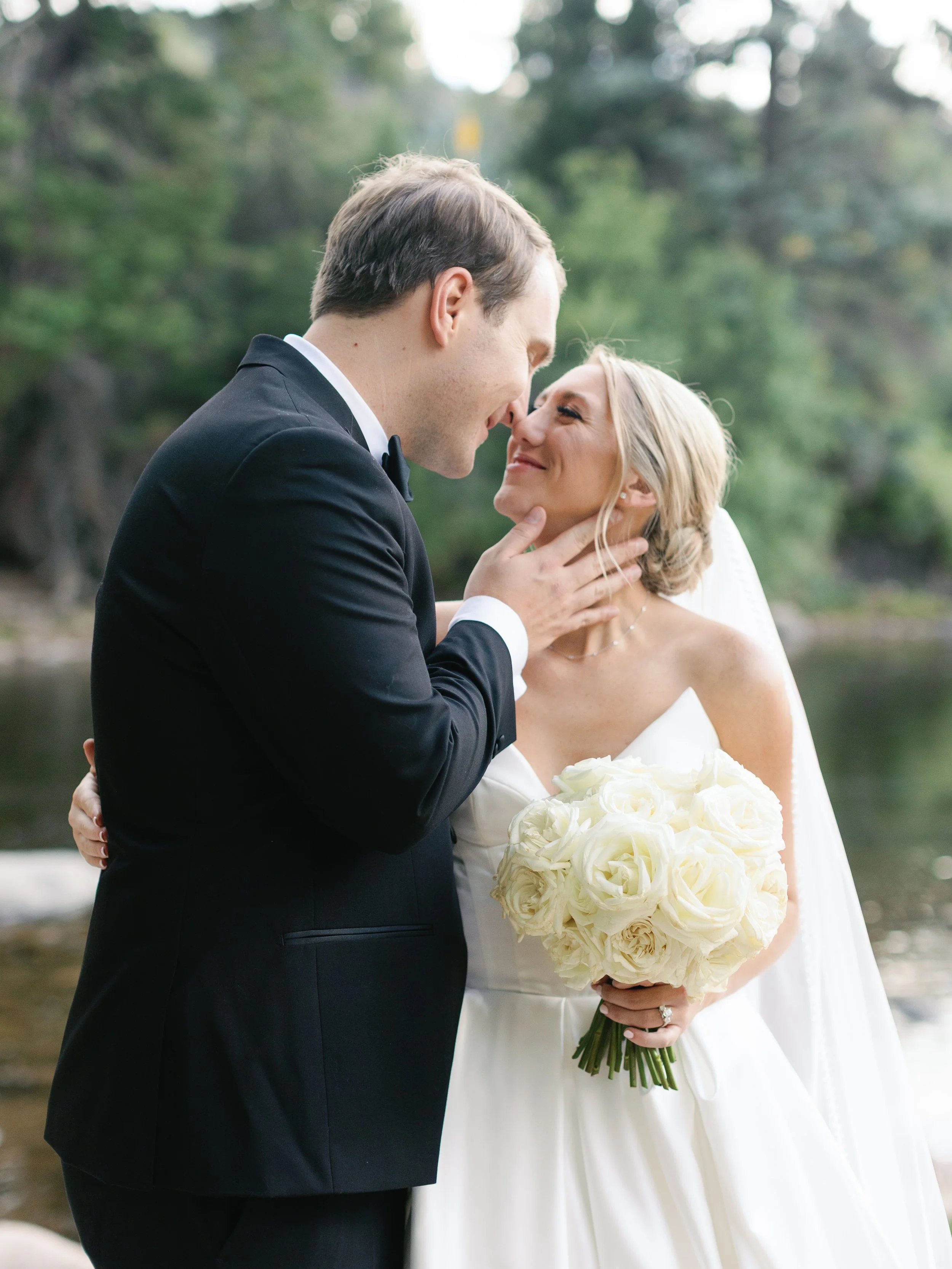 A wedding couple standing close together outdoors, with a forest and river in the background. The groom is holding the bride's face while they look into each other's eyes. The bride is holding a bouquet of white roses.