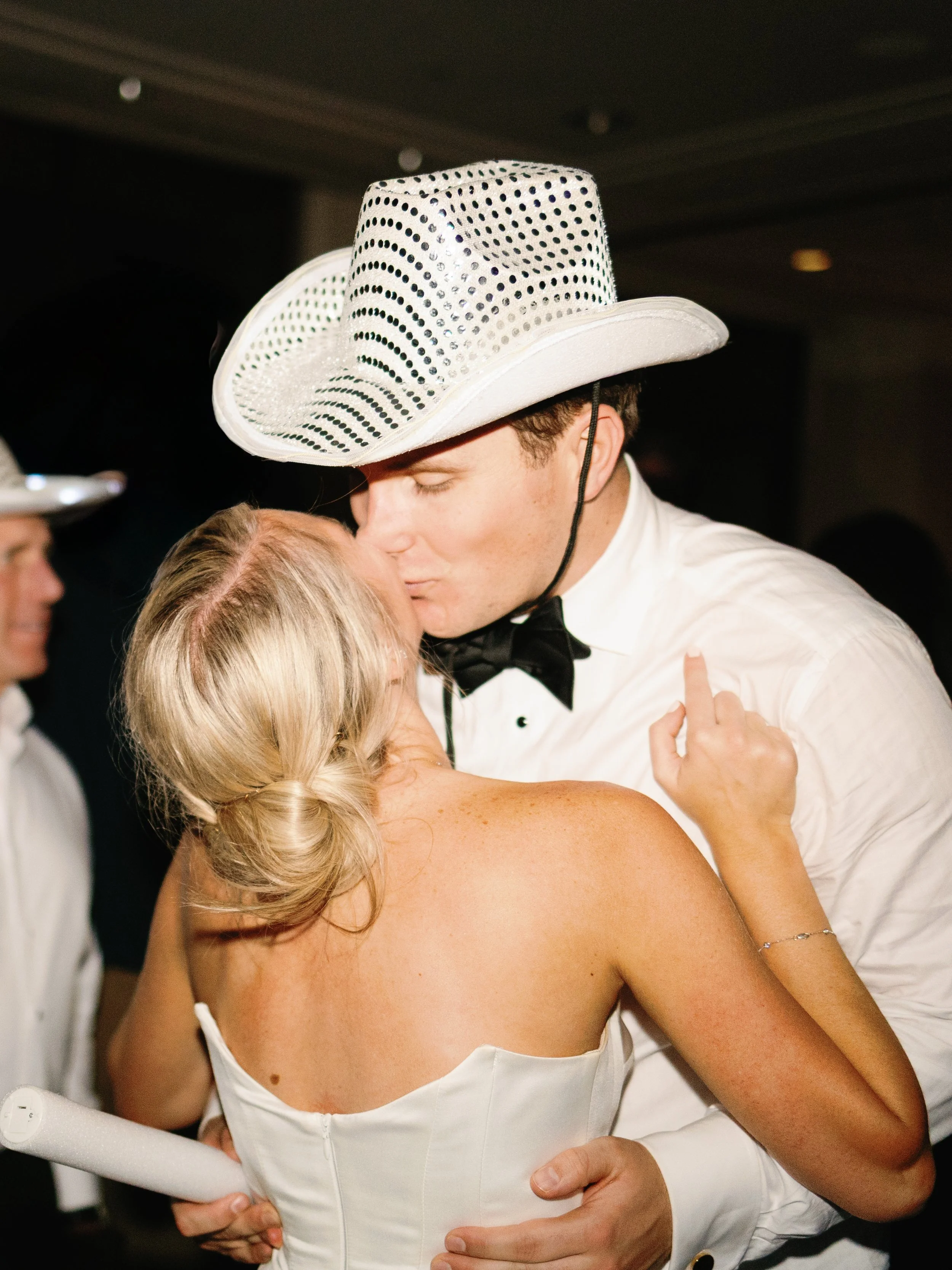 A young woman kissing a man dressed in a white tuxedo and cowboy hat at a party.