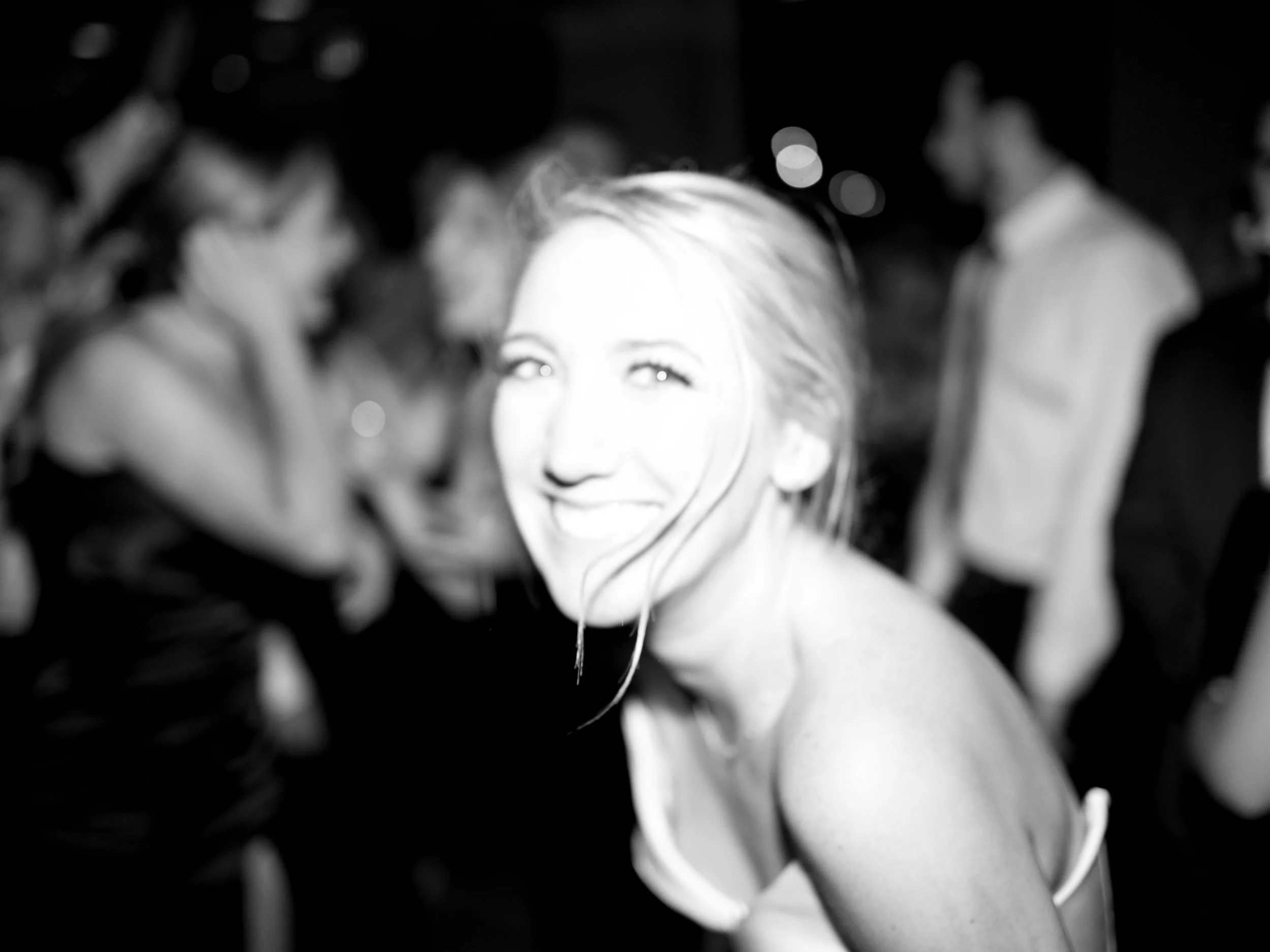 A smiling blonde woman at a social event, surrounded by other people in the background.