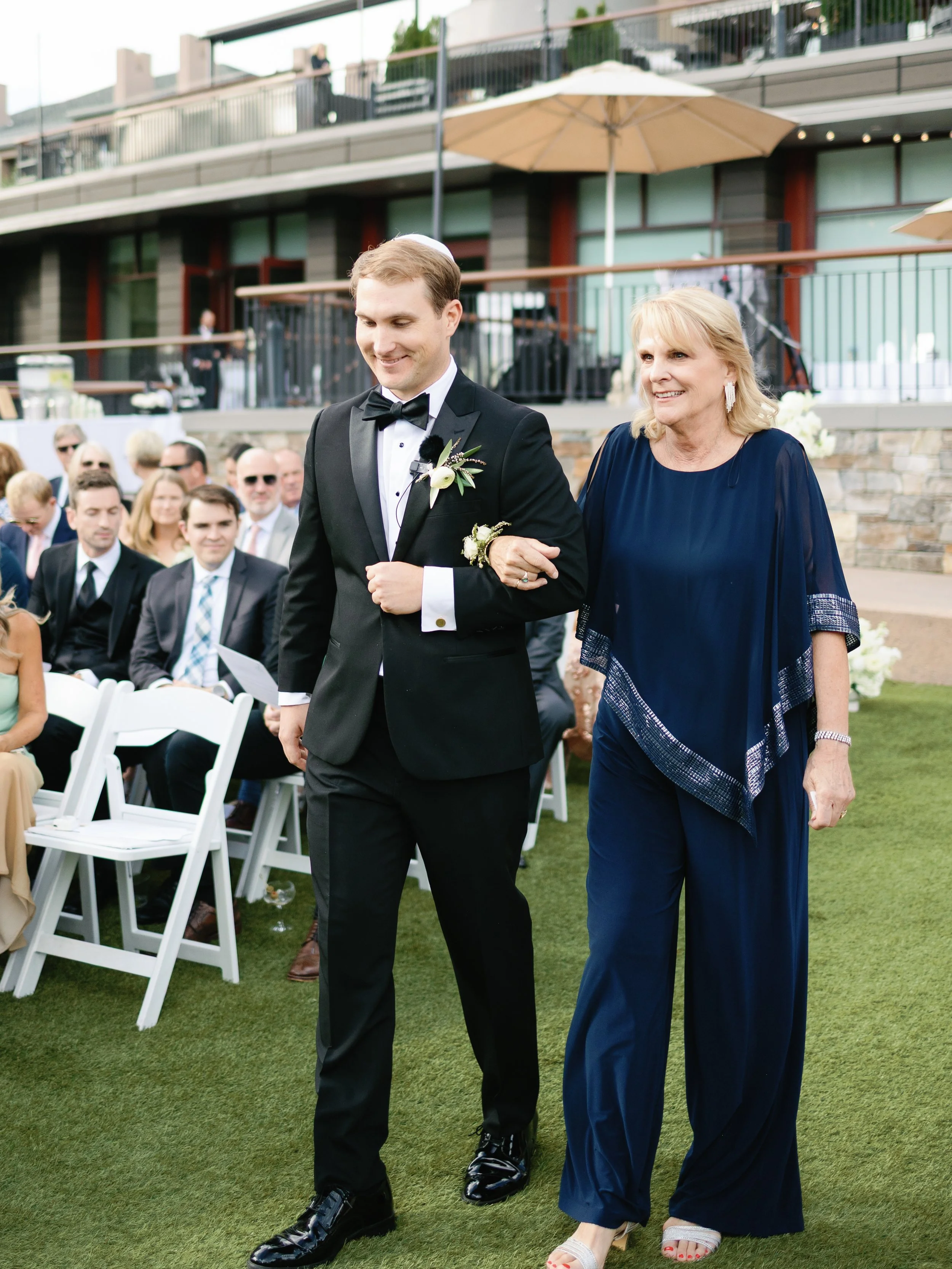 A groom in a black tuxedo walking down the aisle with an older woman in a blue dress at an outdoor wedding ceremony. Guests seated on white chairs in the background, some wearing sunglasses, on a grassy area with a building and umbrellas behind them.