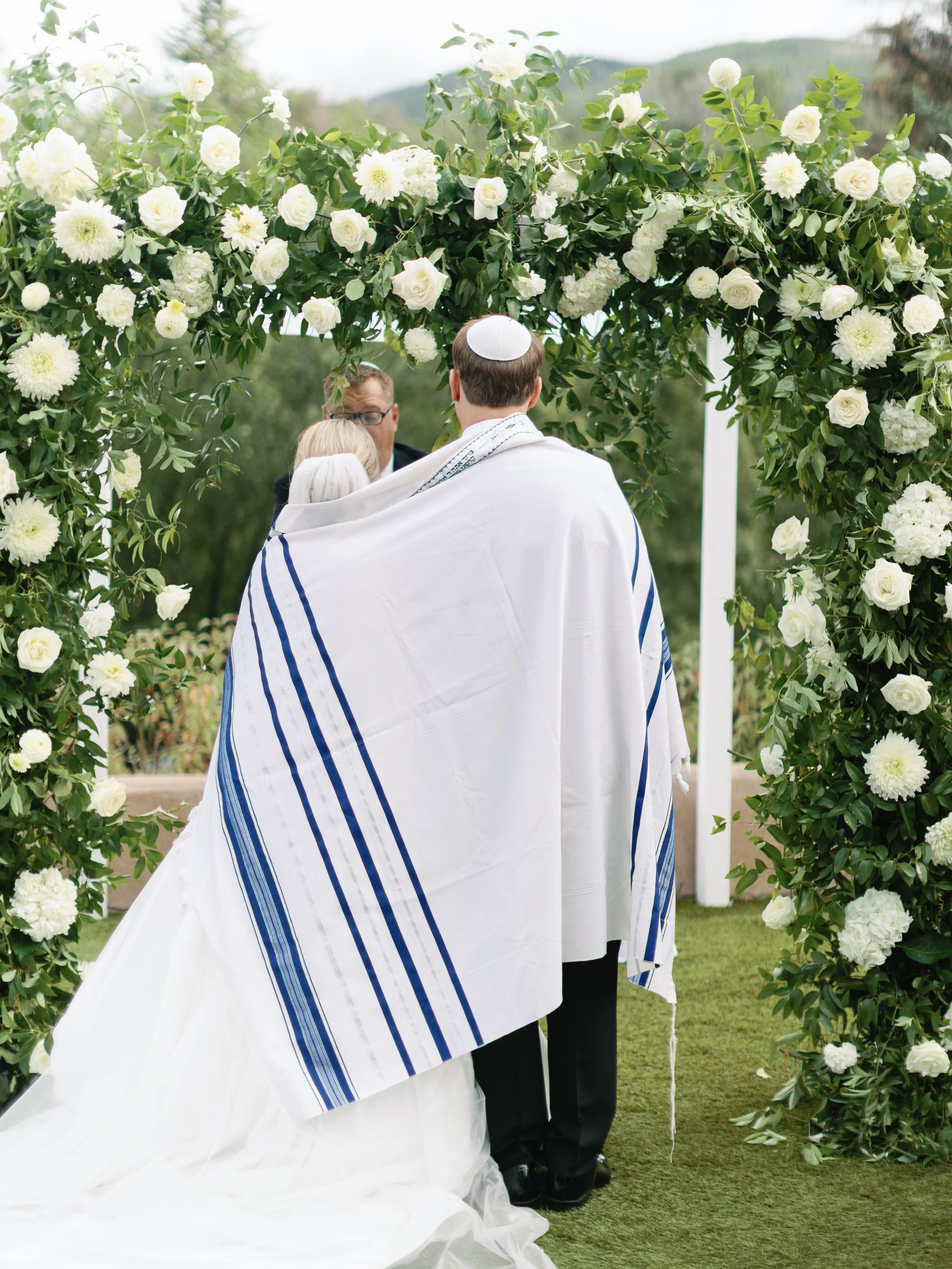 Jewish wedding ceremony with couple under a floral chuppah, with the groom wearing a kippah and wrap, and the bride in a white dress, during daytime outdoors.