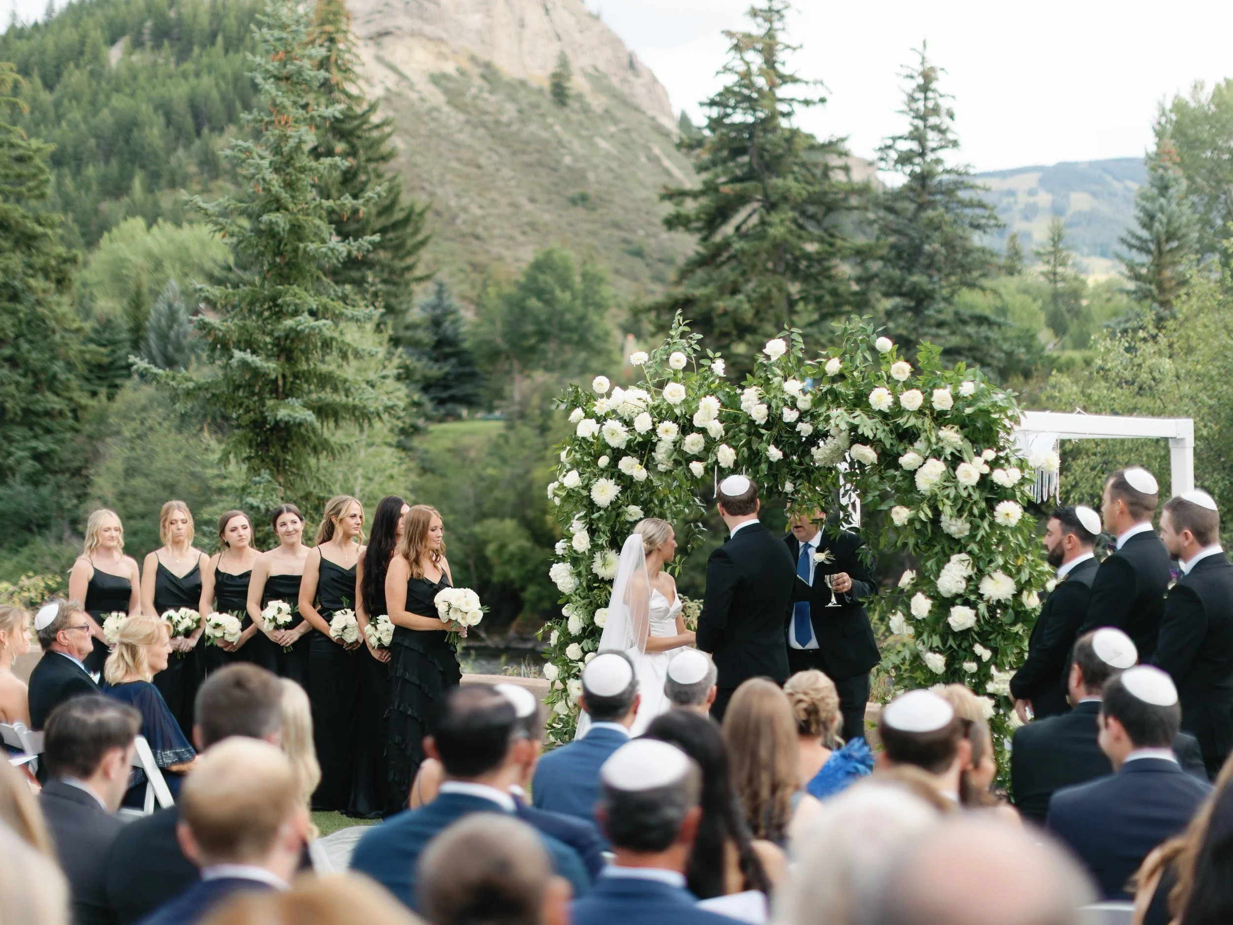 Outdoor wedding ceremony with a couple exchanging vows under a floral arch, surrounded by bridesmaids in black dresses and groomsmen in dark suits, with guests in formal attire seated nearby, set against a backdrop of trees and mountains.