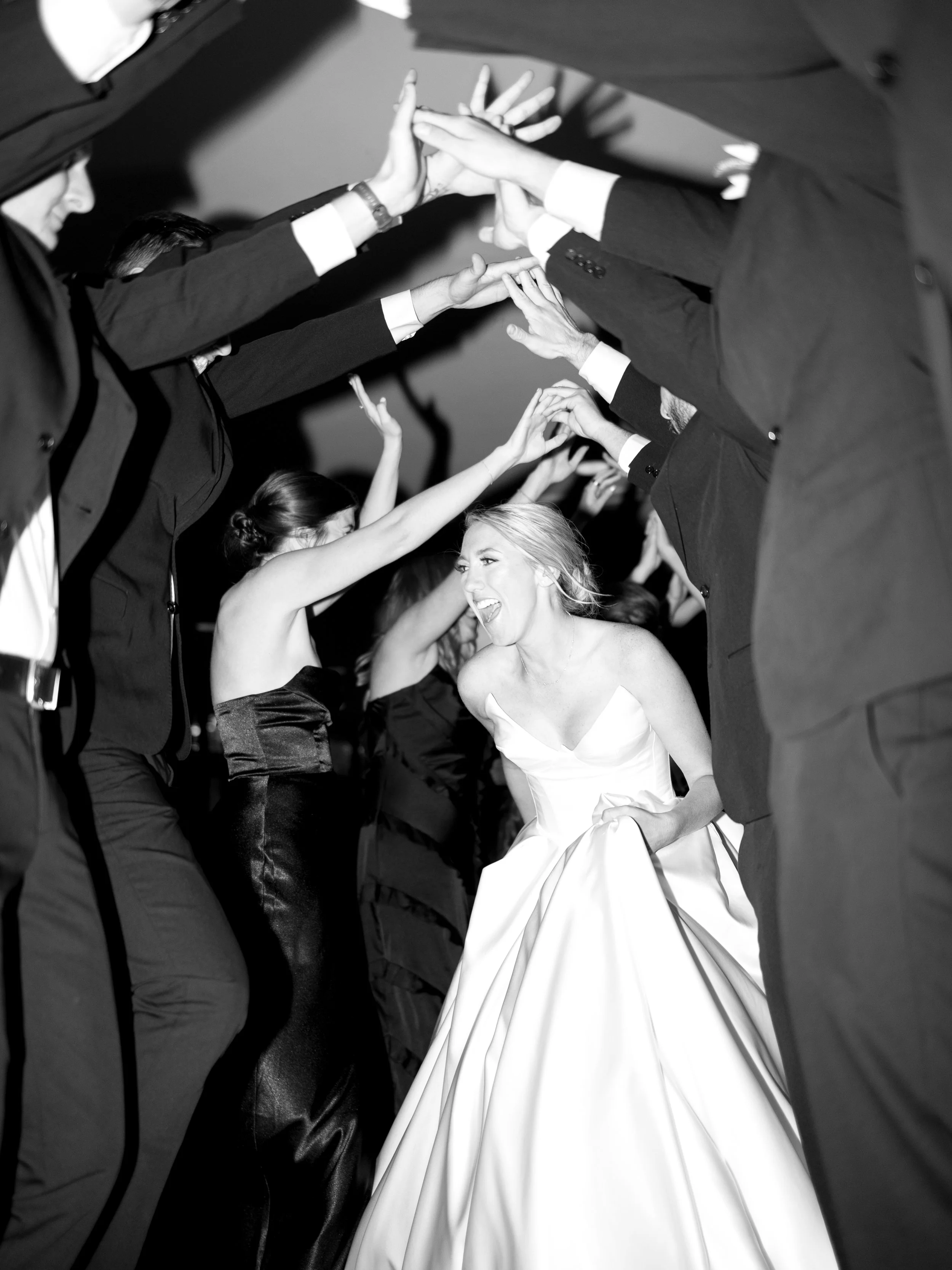 The bride in a white wedding gown smiling and laughing as she walks through a tunnel of guests dressed in formal attire, all reaching out and holding hands above her head.