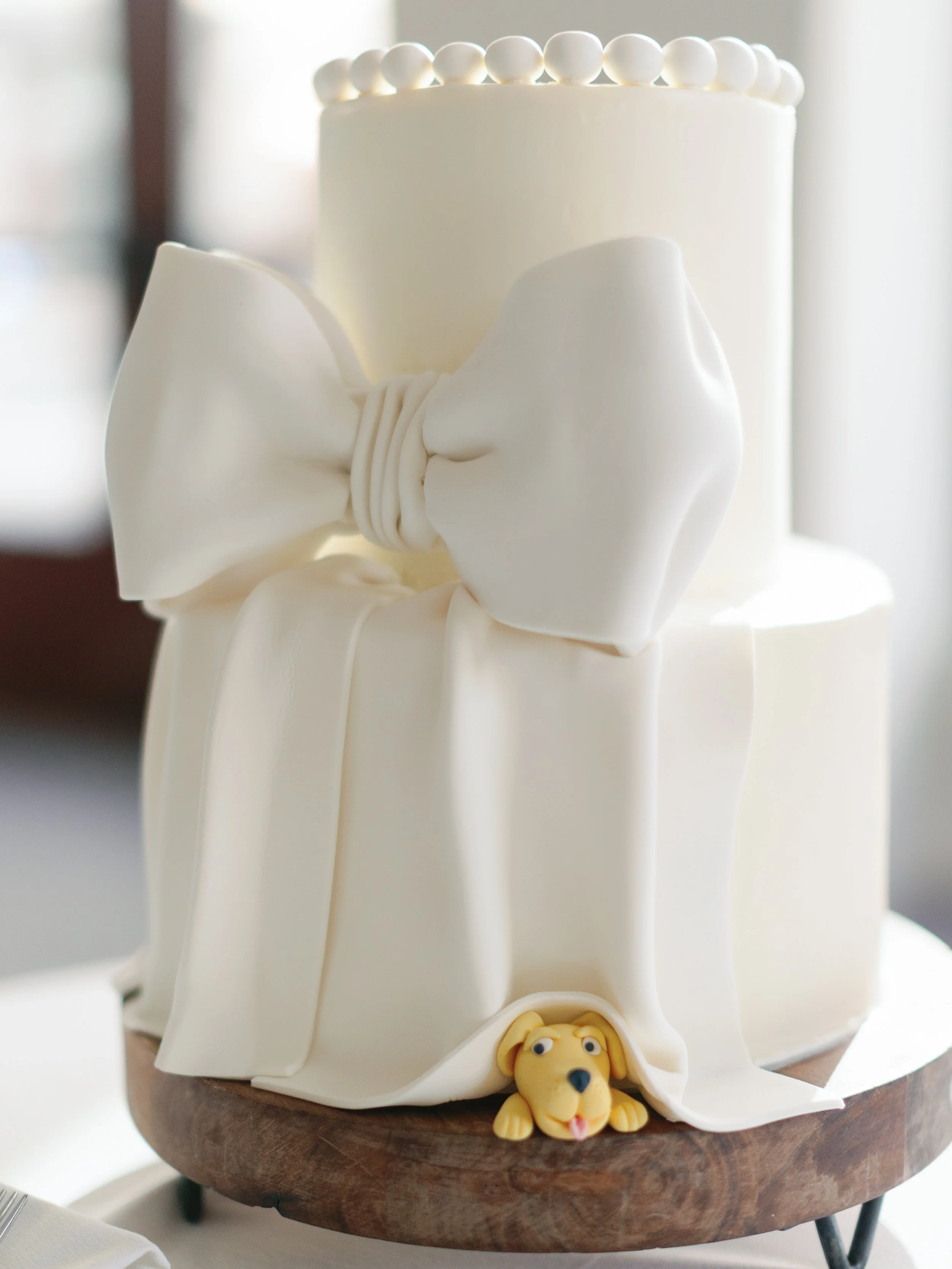 Two-tiered white cake with a large white bow decoration and small white round accents on top; a small yellow dog figure at the base of the cake.